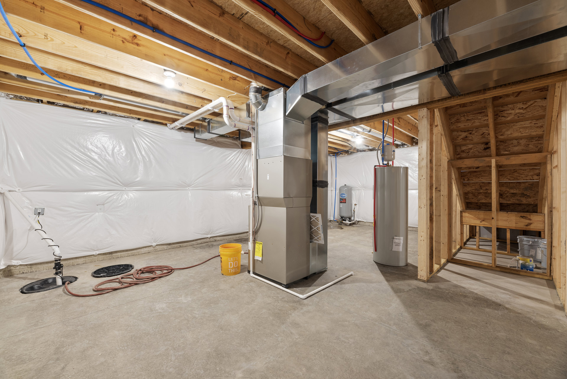 Basement room with exposed beams, plaster walls, composite flooring, large metal HVAC unit, yellow bucket with white text, white door, grey cylinder with blue and red sticker, and