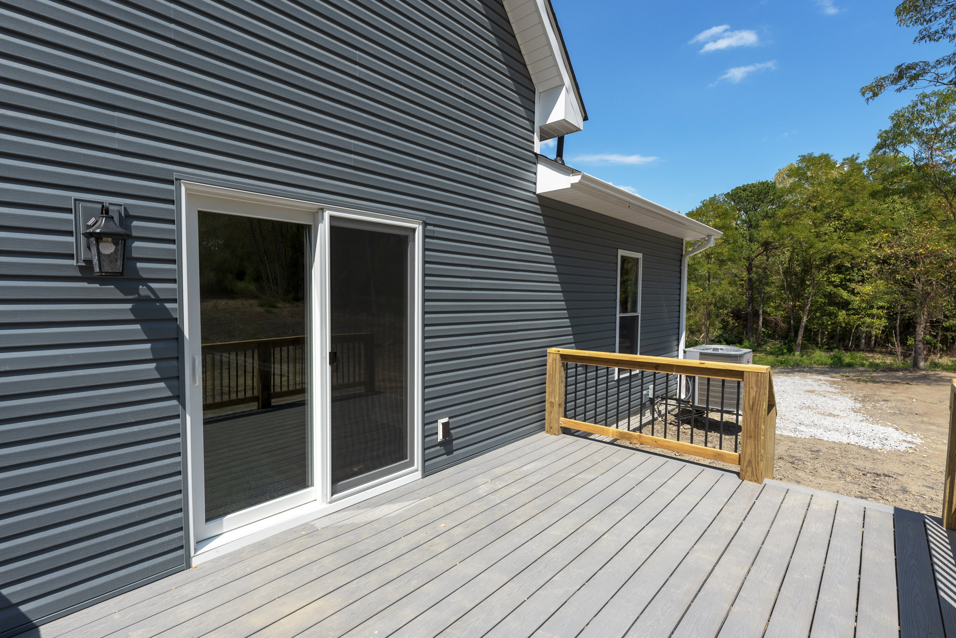 Wooden deck with metal railing, sliding glass door, and exterior siding; trees and sky visible in background.