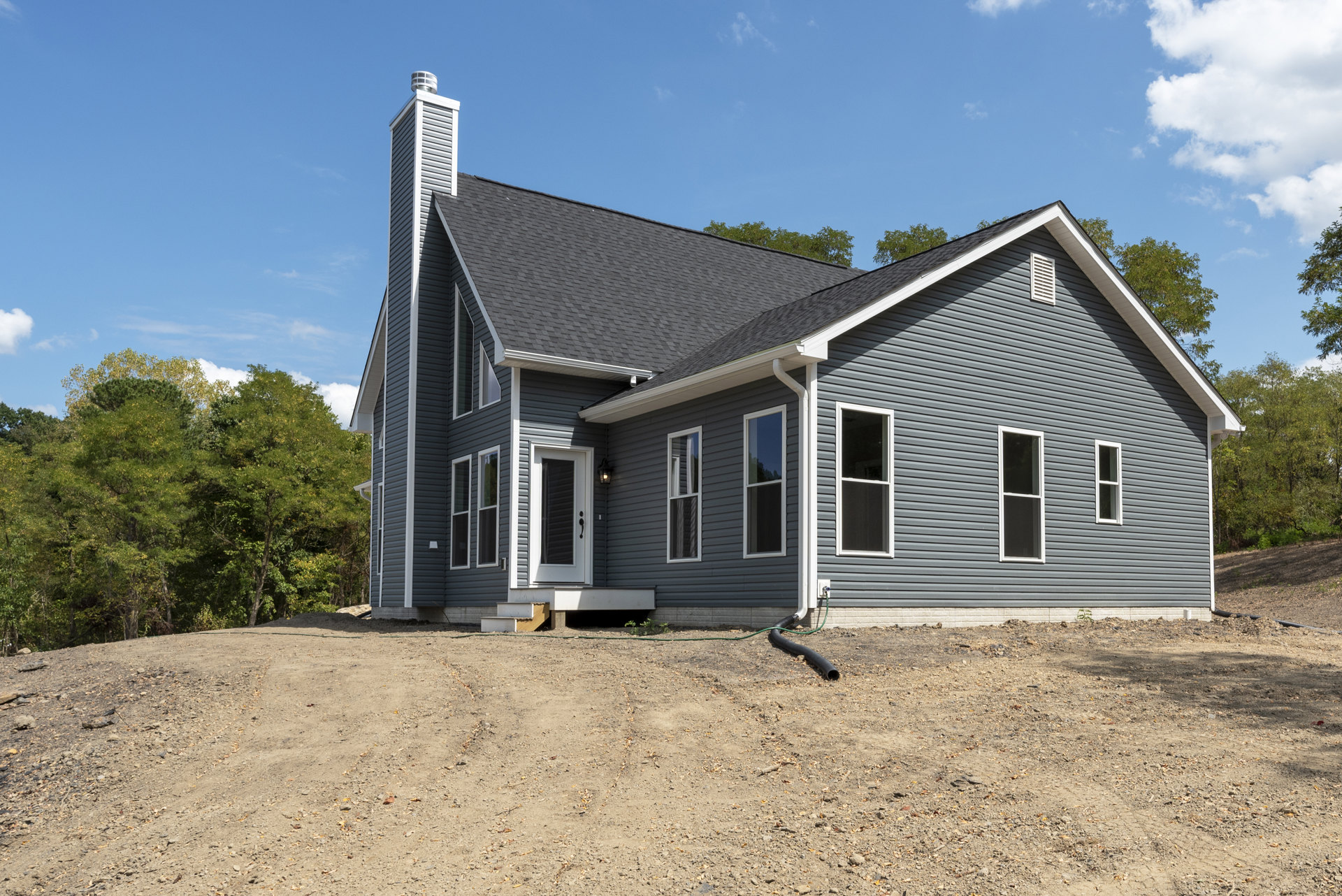 Two-story home with white-framed windows and white doors, set against a dirt hill with surrounding trees and a garden hose extending from the exterior wall.