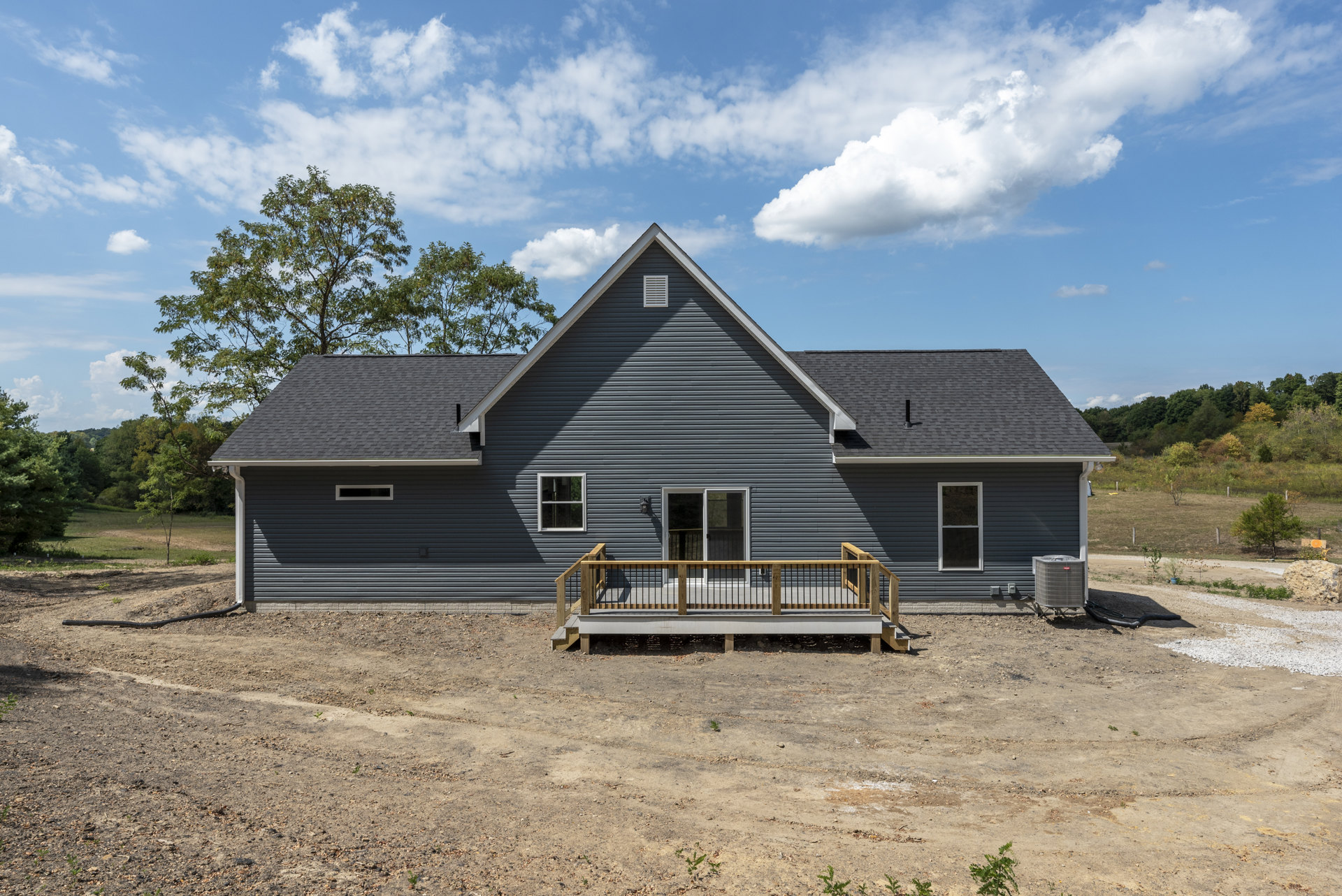 Partially built house with exposed wooden deck and railing, large window, gray square heater unit, roof vent, surrounded by trees and cloudy sky.