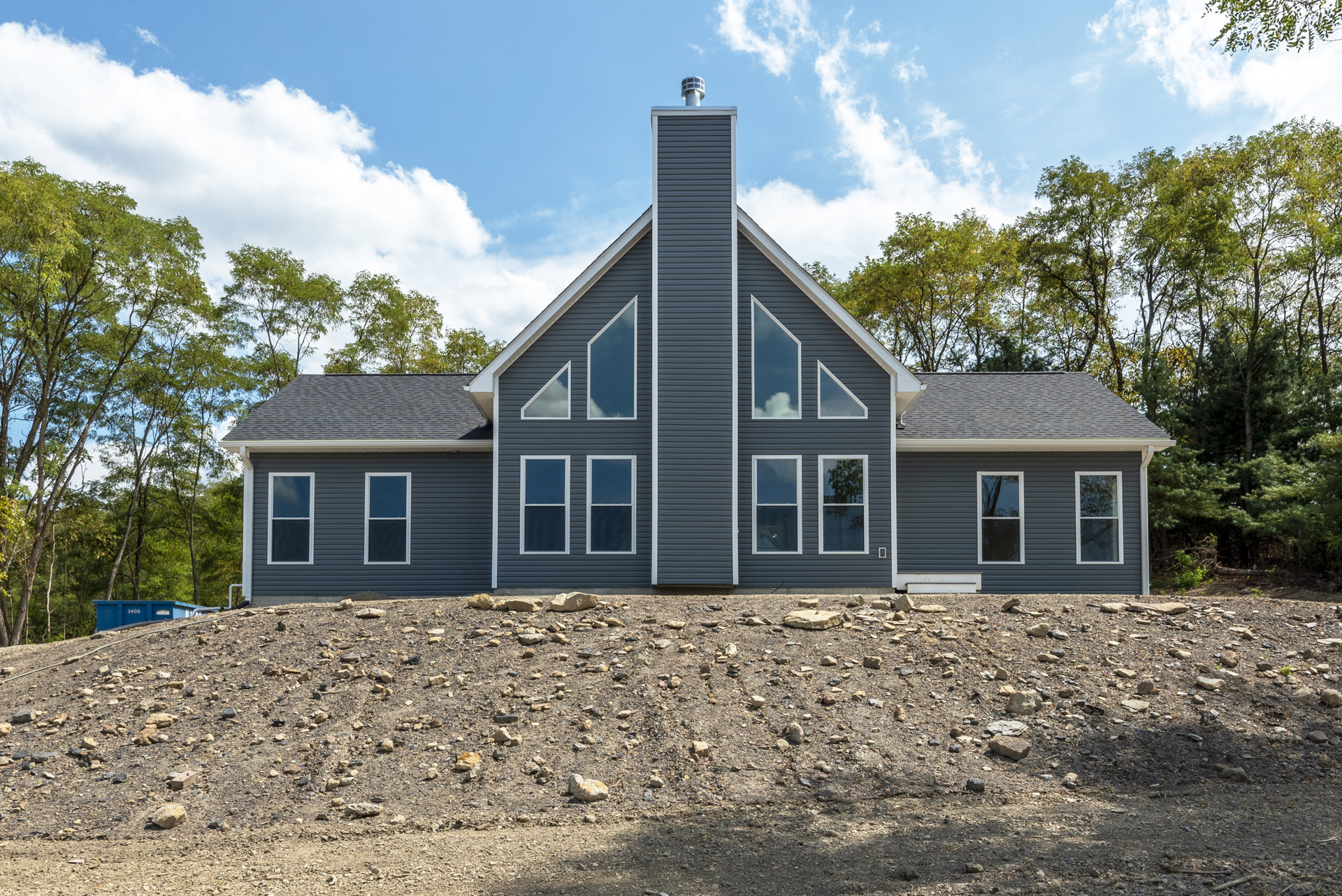 Two-story house with light siding, side chimney, white-framed windows reflecting trees, rocky ground in front, cloudy sky overhead