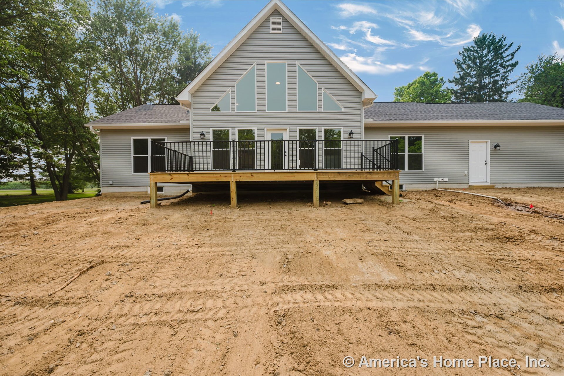 Elevated wooden deck with black metal railings, large geometric windows, gray vinyl siding, gable roof, exterior wall-mounted lighting, and crisp white trim on a newly constructed