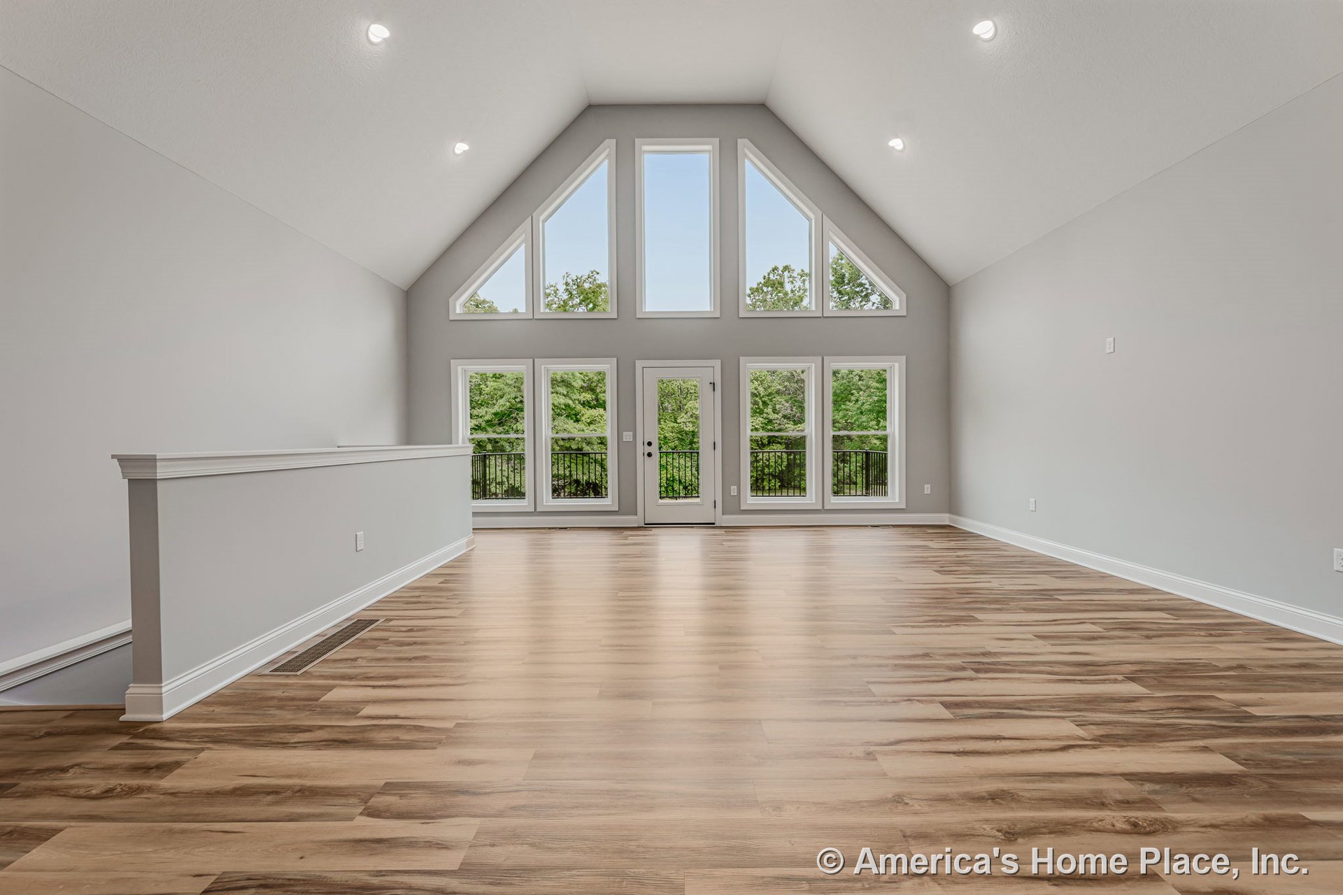 Vaulted ceiling living room with floor-to-ceiling windows and a central glass door opening to a balcony; light wood plank flooring, white trim and baseboards, and abundant natural