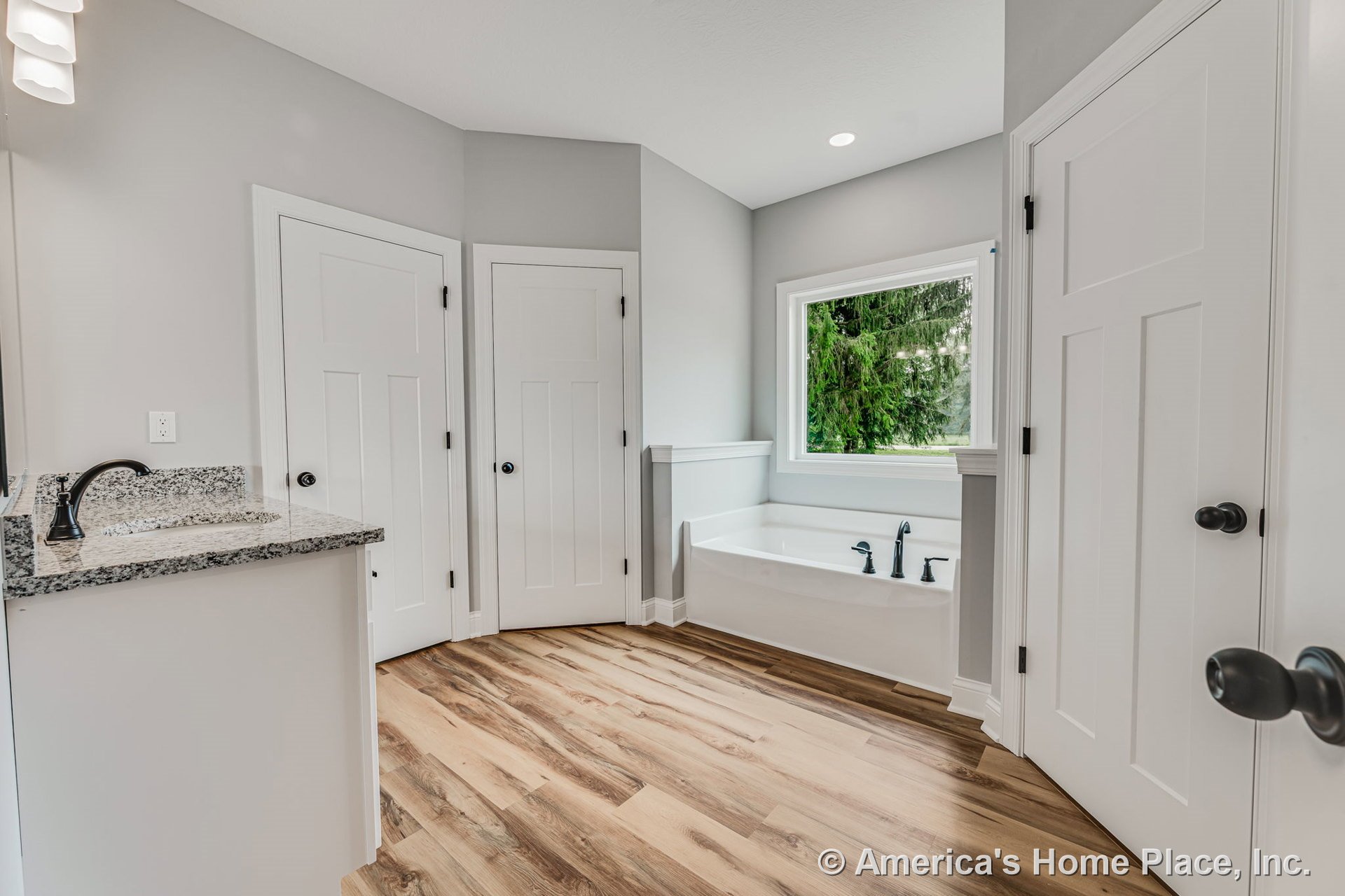 Soaking tub with black fixtures positioned beneath a large window, granite vanity countertop with white cabinetry, wood-look plank flooring, white paneled doors, recessed ceiling