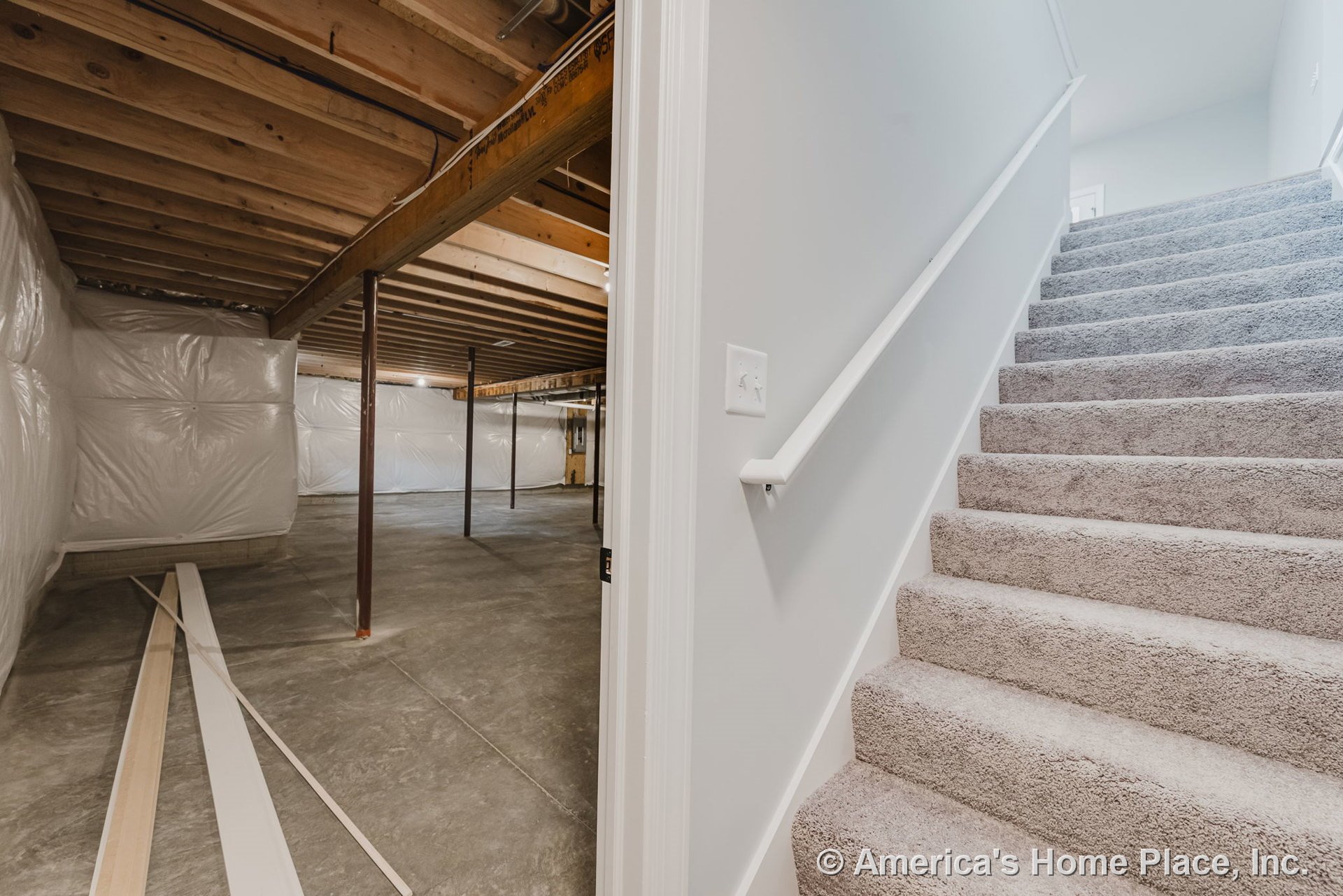 Carpeted staircase with white painted railing descends into unfinished basement featuring exposed wood ceiling joists, concrete flooring, and plastic-wrapped walls.