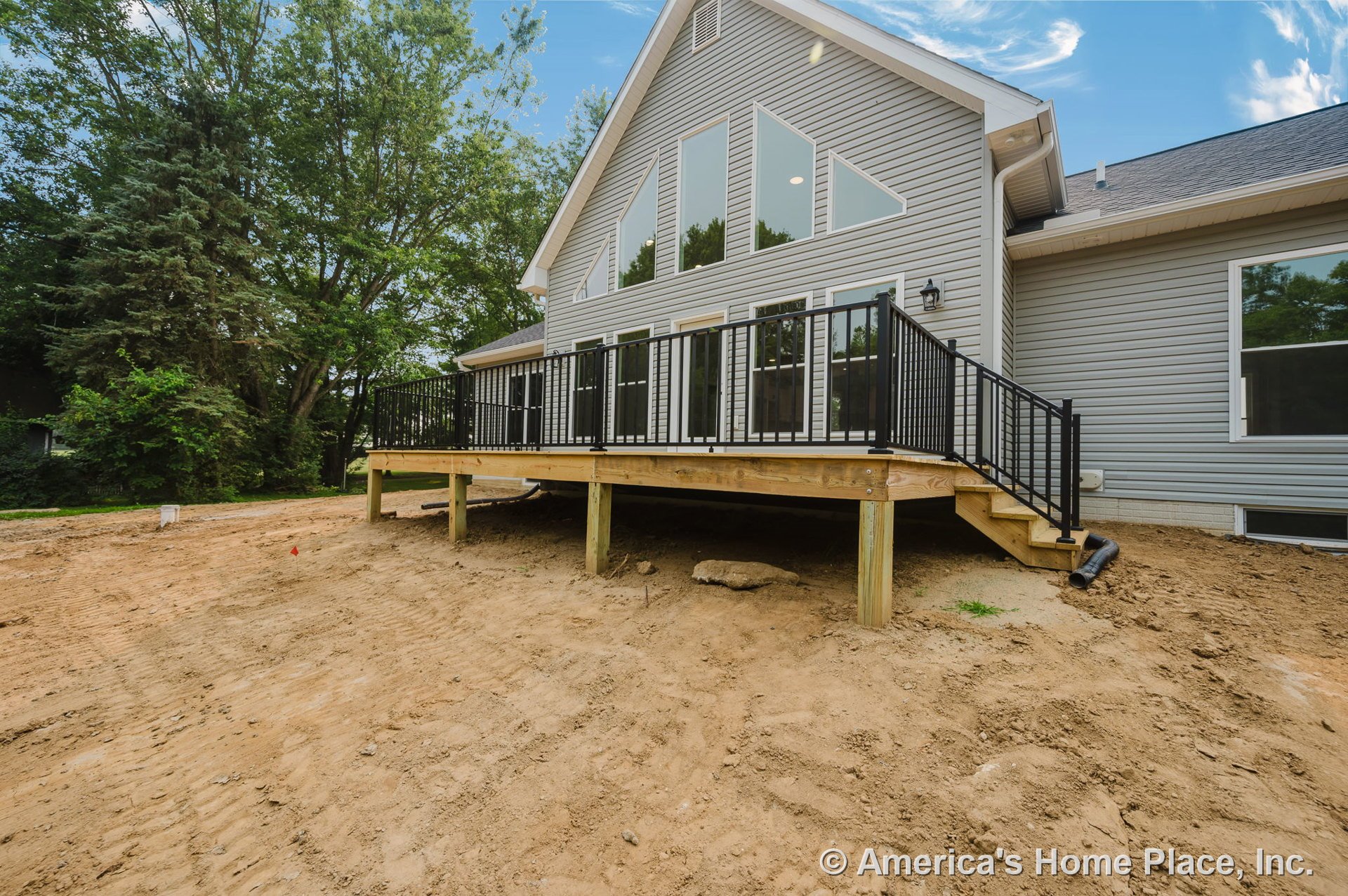 Elevated wooden deck with black metal railings, large triangular windows, gray vinyl siding, outdoor staircase, and unfinished yard.