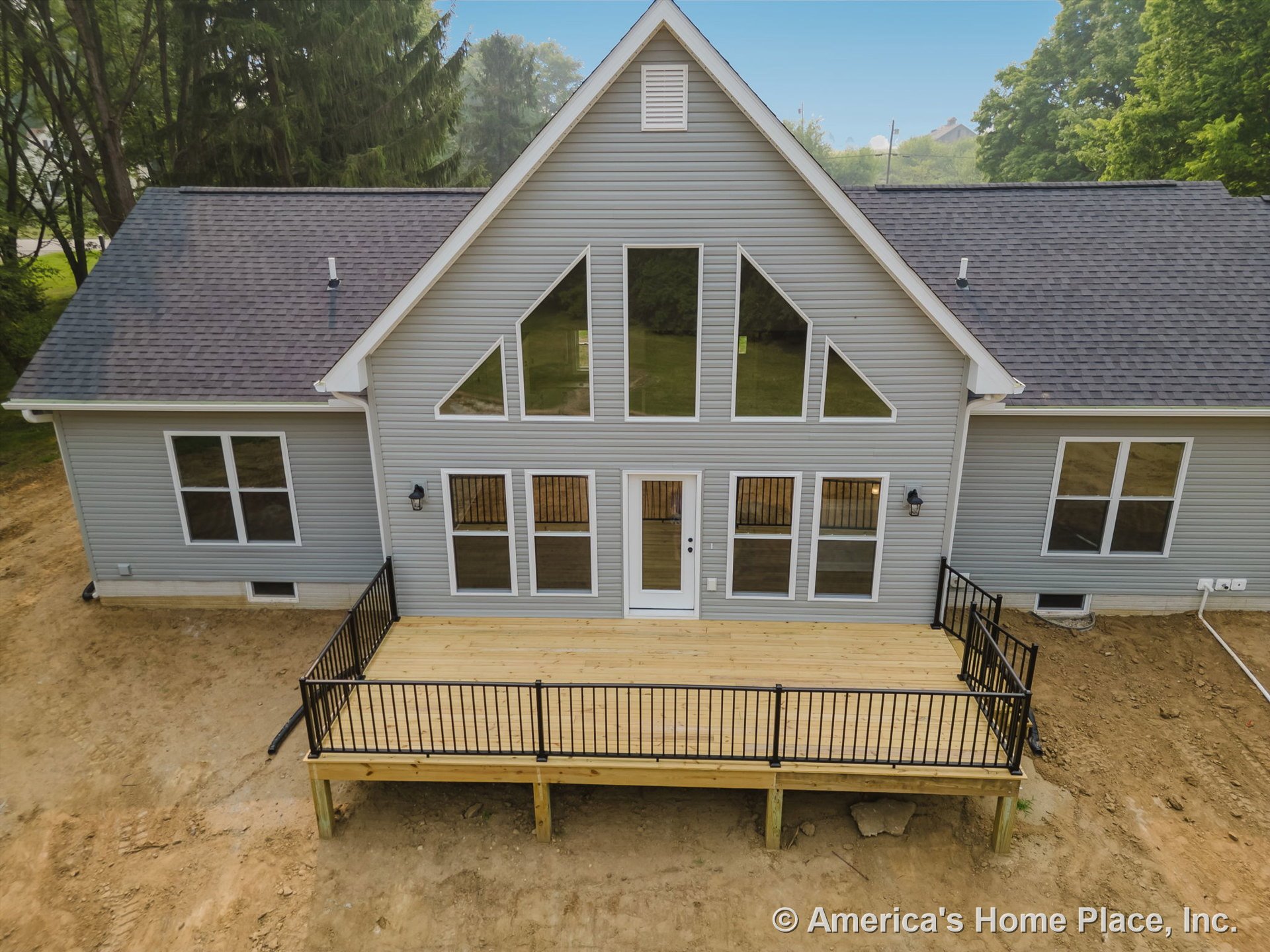 Spacious wooden deck with black metal railing, gray vinyl siding, geometric windows, gable roof, exterior wall lights, and white trim surrounding entry door.