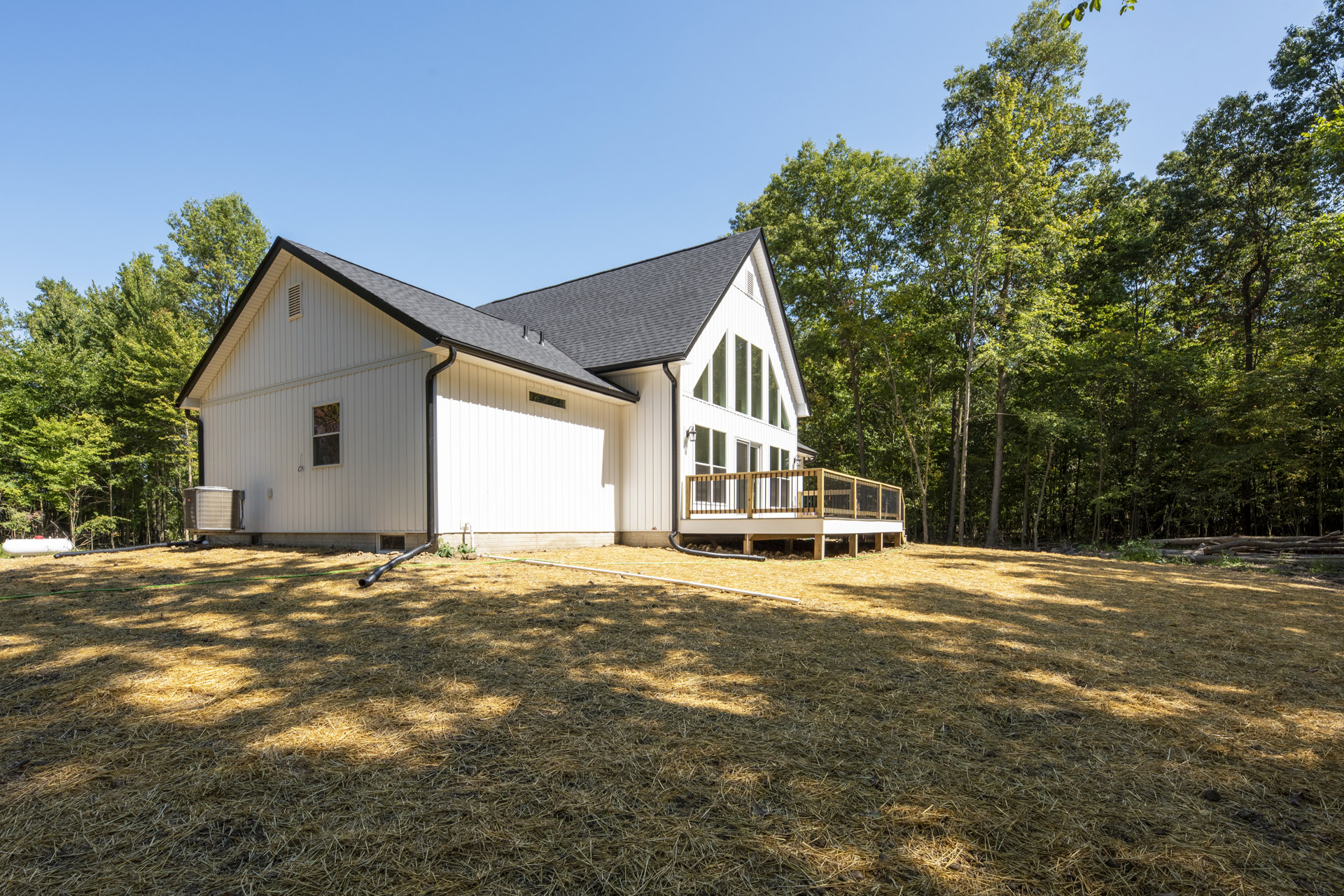 White siding house with wooden deck featuring black railing, surrounded by straw-covered yard and mature trees in the background, large air conditioner visible on roof.
