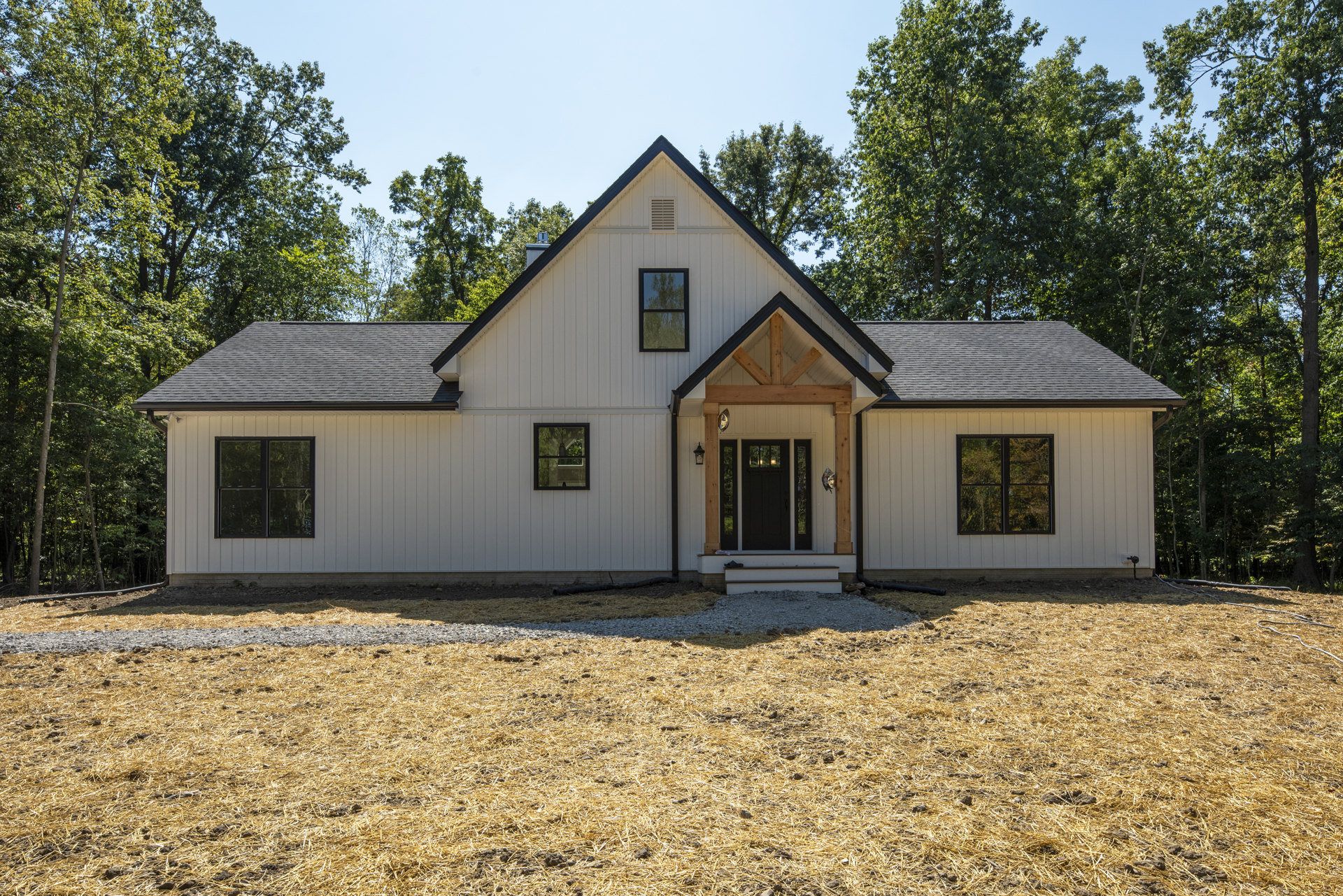 White siding house with black roof, black framed windows, glass panel black door with wooden trim, tree reflected in window, illuminated window, front yard with grass, rocks, and