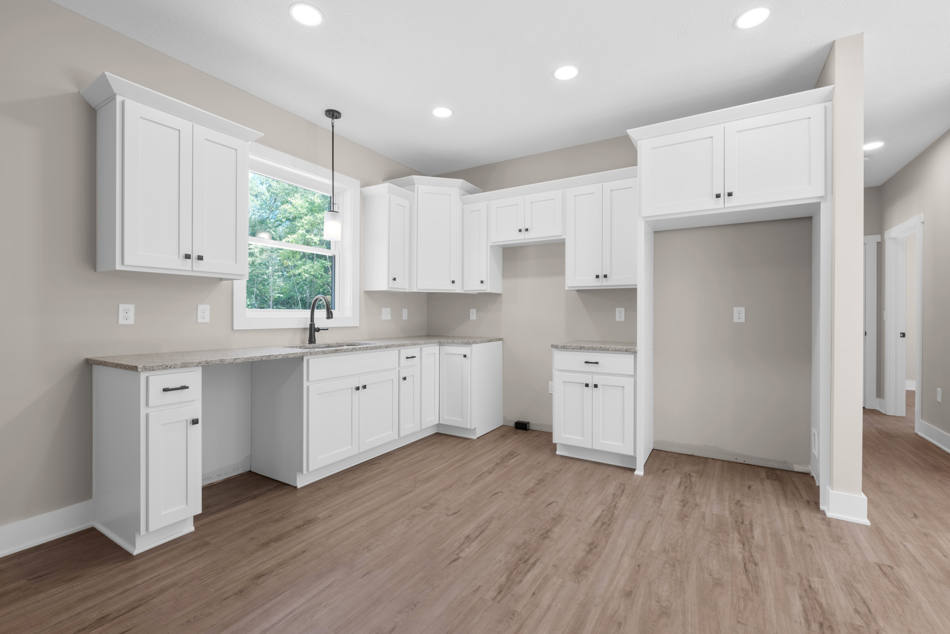White kitchen cabinets with black handles, wood plank flooring, black gooseneck faucet at the sink, large window overlooking trees, light countertops, and drawers.