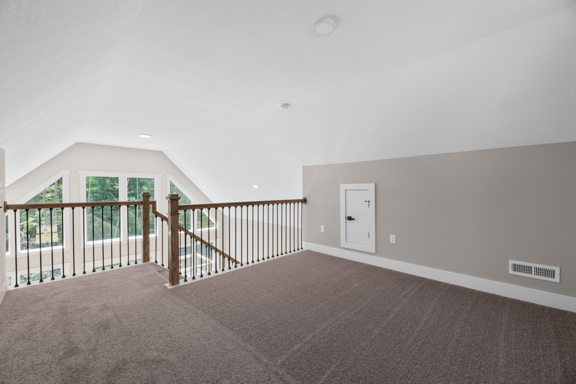 Staircase with wooden railing, white door with black handle, carpeted floor, white wall vent, large windows showing trees outside
