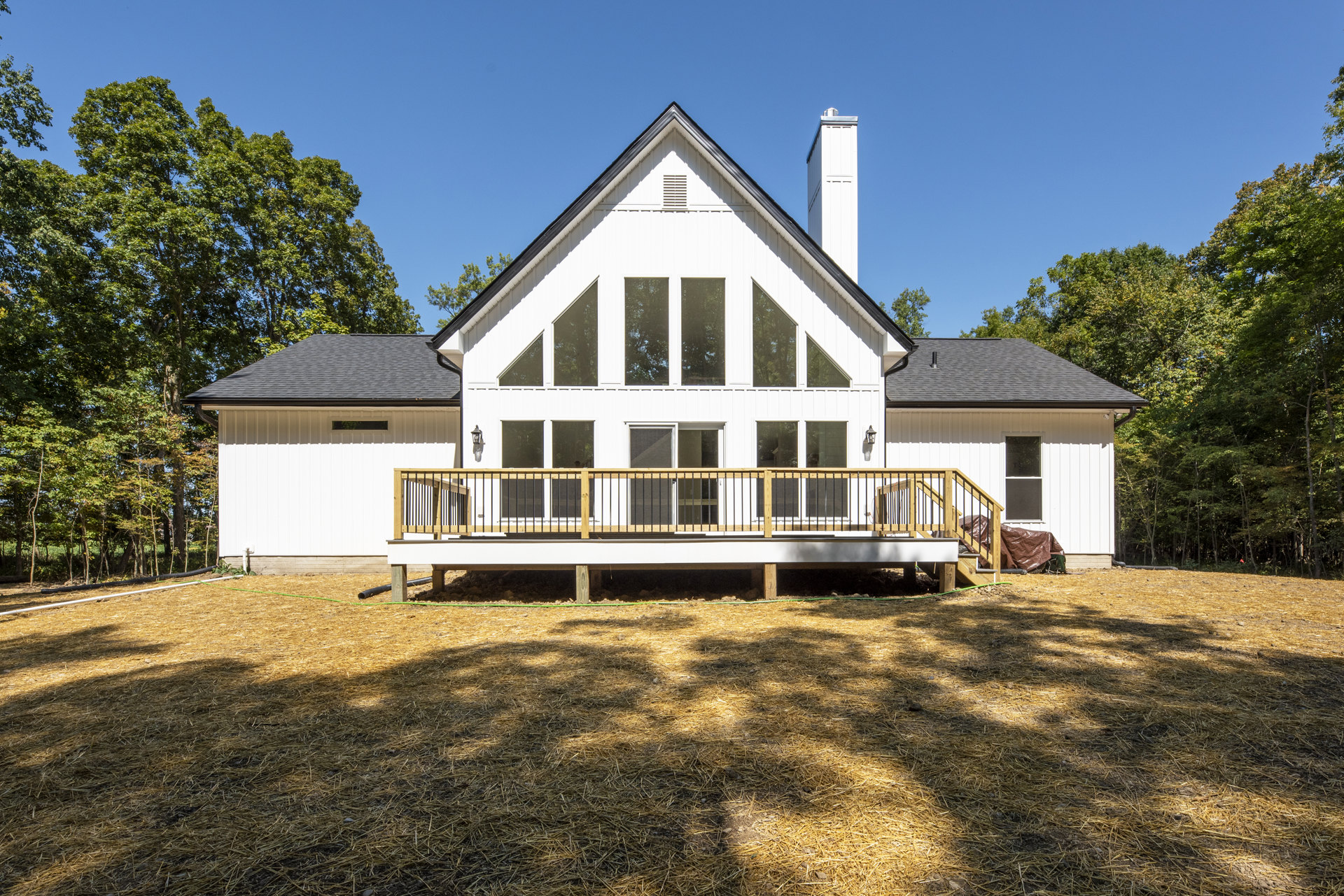Two-story home with wood deck and covered porch, white siding, brick chimney, wooden railings, windows, and surrounding trees