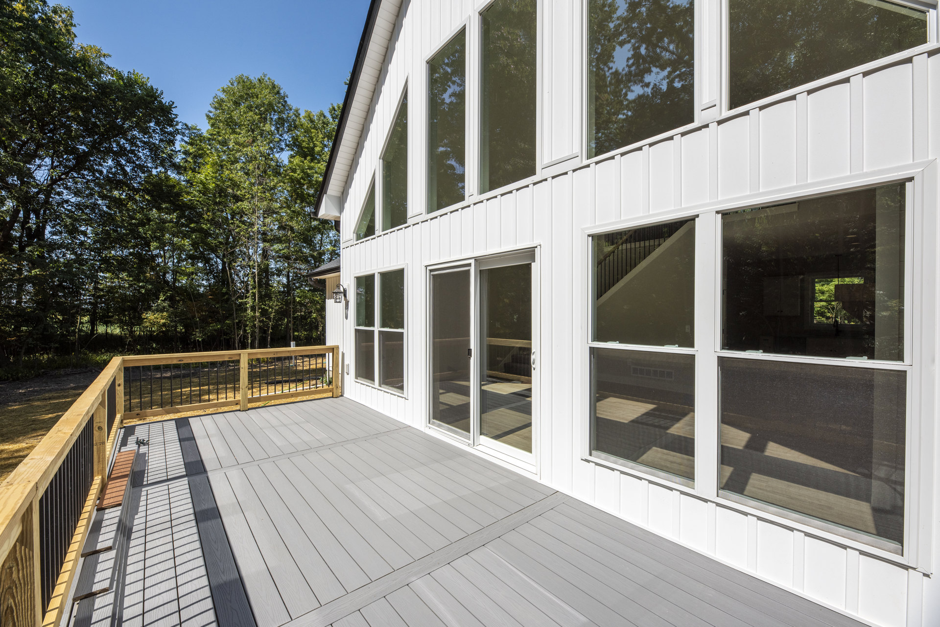 Spacious white house with multiple windows, wooden deck featuring metal railing, glass doors, bench, and surrounding trees