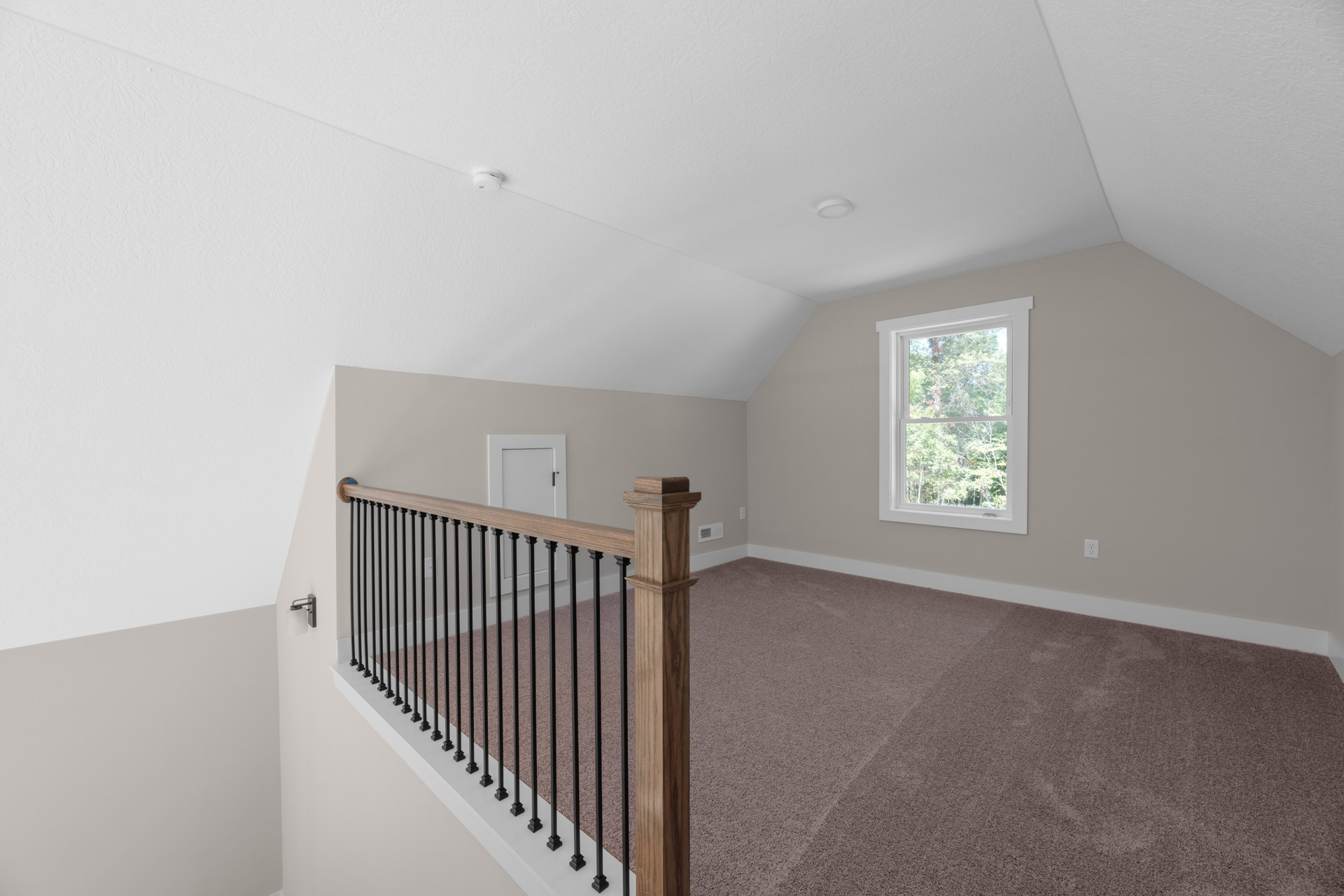 Open staircase with black metal railing, light wood steps, large window showing trees outside, white door with black handle, plaster walls, and hardwood flooring.