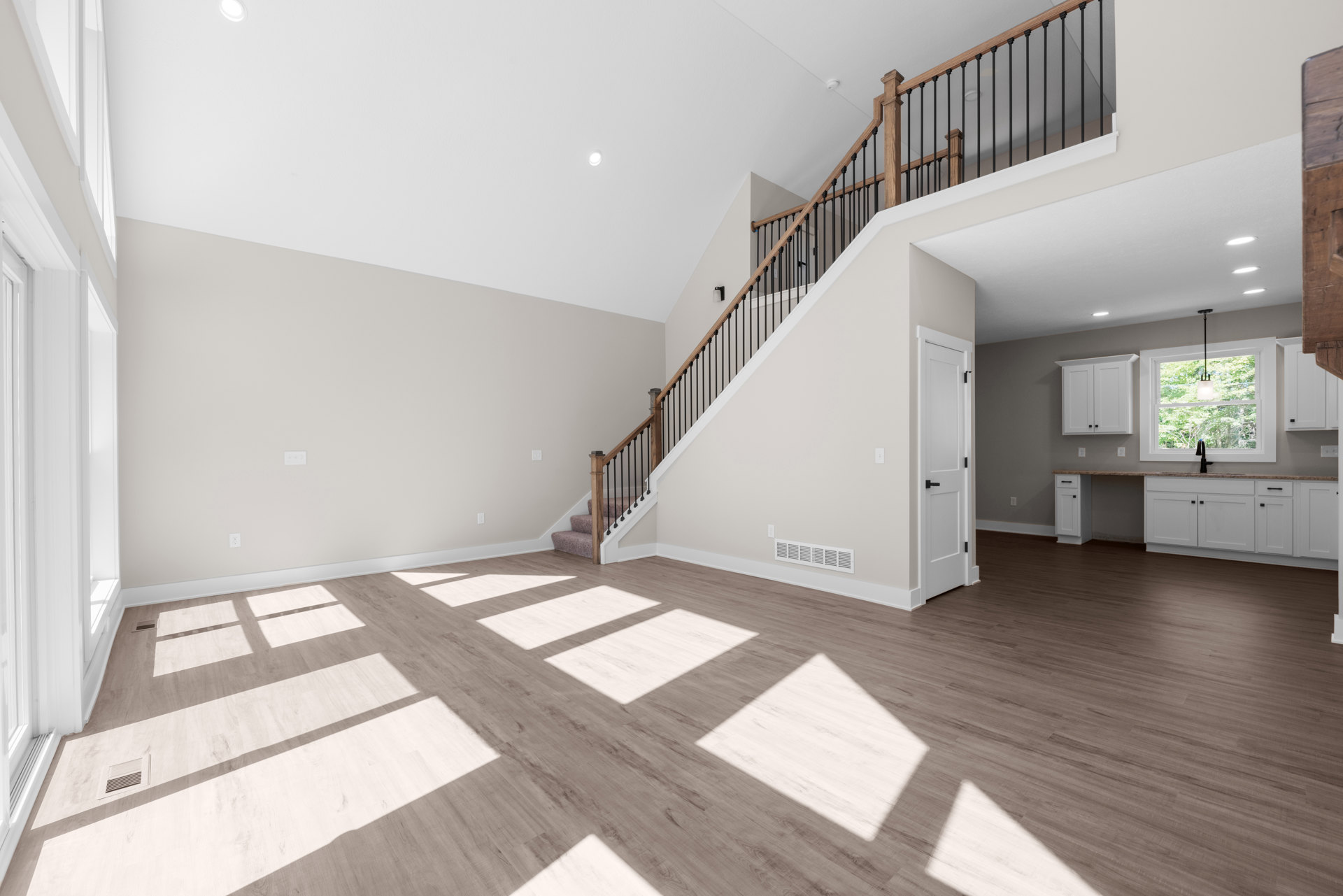 Open living area featuring wood flooring, white plaster walls, central staircase with dark railing, large windows allowing natural light, white door with black handle, ceiling