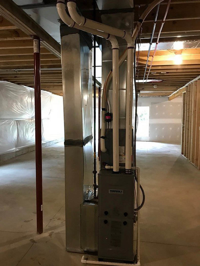 Exposed metal pipes and machinery in an unfinished basement with steel beams, white wall coverings, and a column wrapped in tape