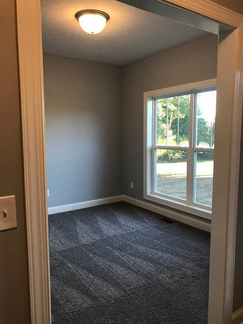 Neutral-toned carpeted room with a large window overlooking green trees, white walls, ceiling light fixture, and wood trim.