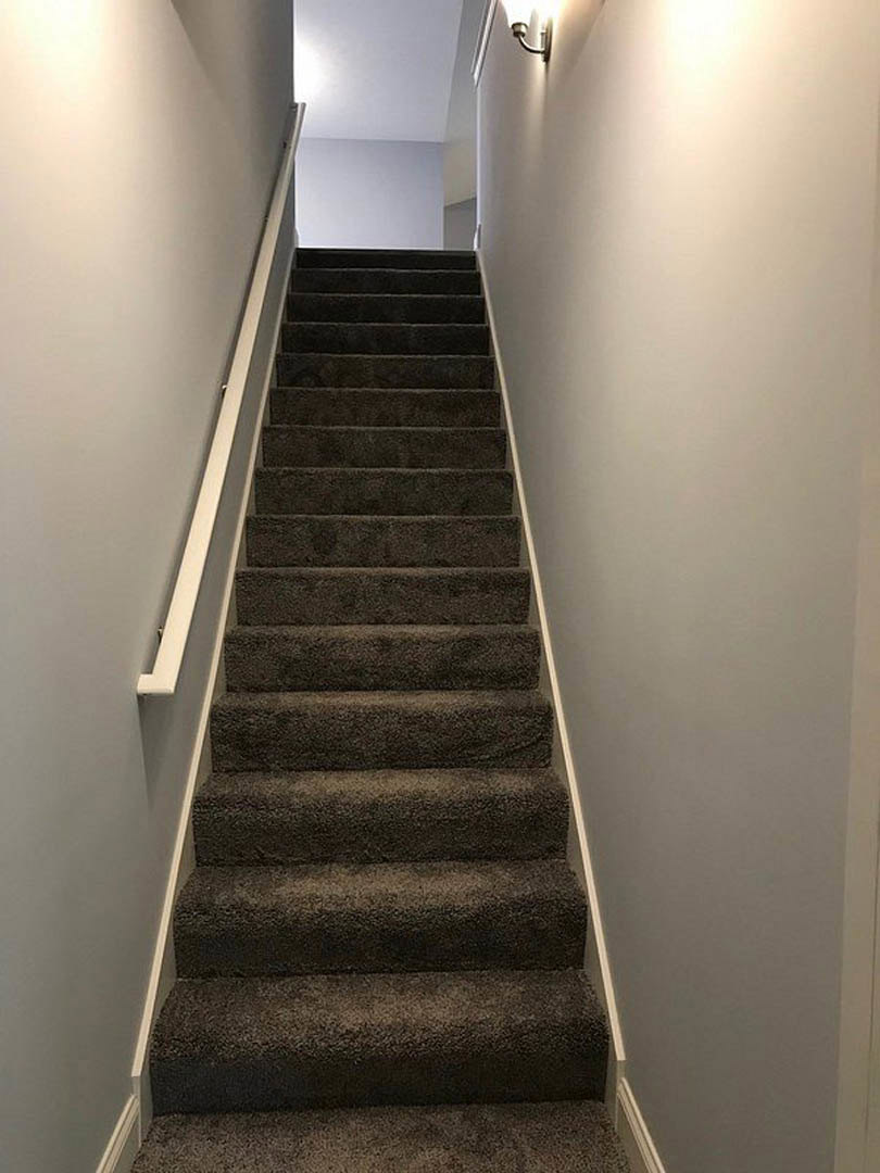 Carpeted staircase with wooden handrail, white walls, and ceiling-mounted light fixture illuminating the space