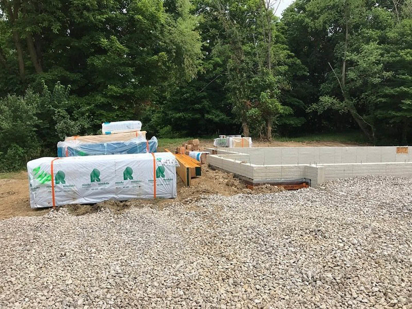 Construction site with stacked bricks, gravel ground, metal fence, white tarp with green logos, partially built white brick wall, scattered rocks, and exposed wood beam