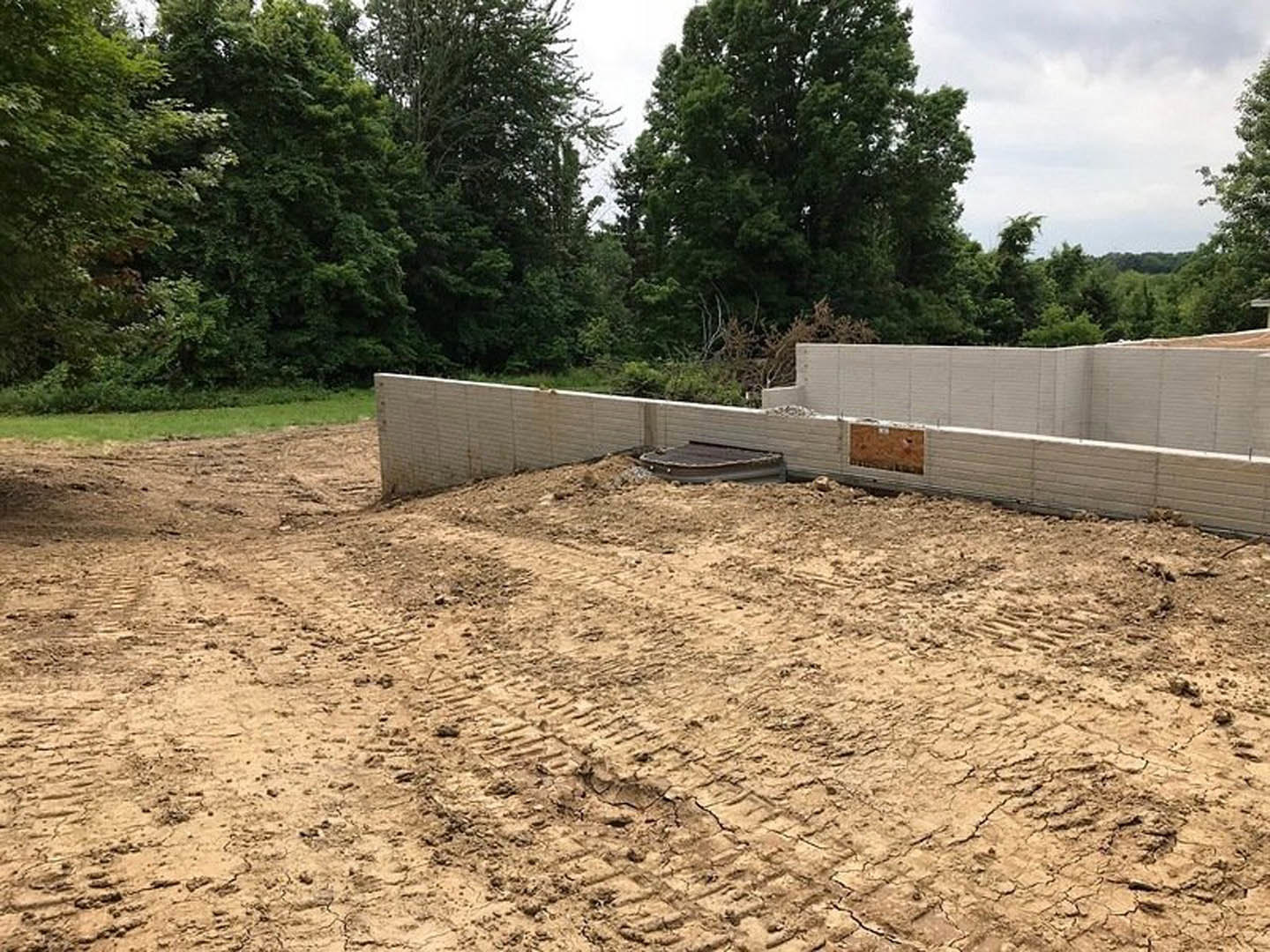 Dirt field with tire tracks, partially constructed wall, worker on site, tree-lined background, unfinished building, exposed soil and materials