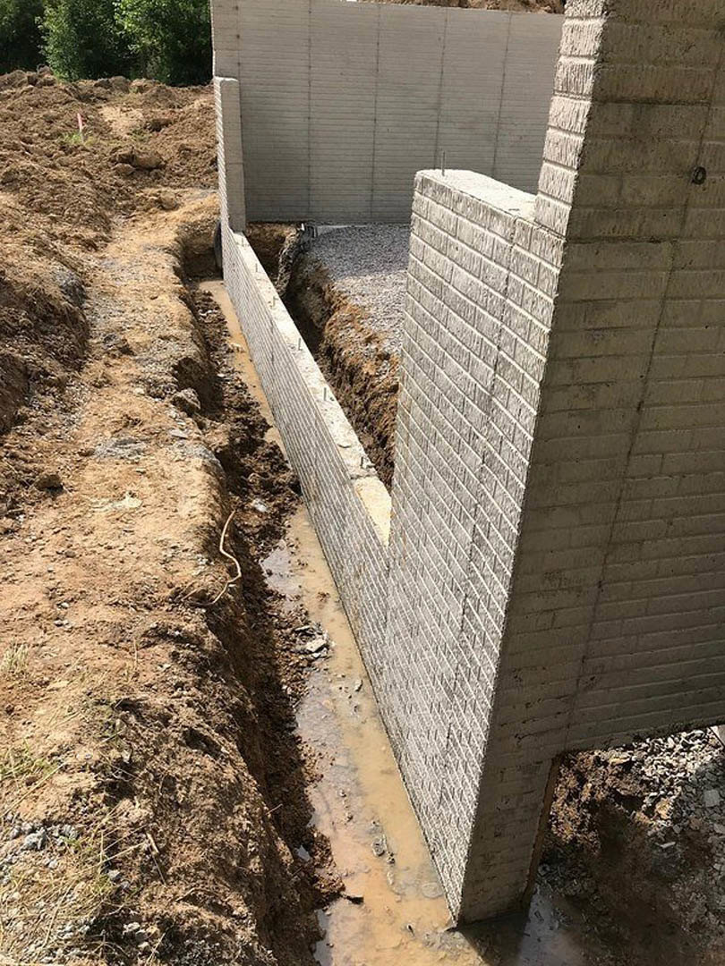 Concrete exterior wall with circular hole in the ground, surrounded by soil, rocks, and sparse vegetation