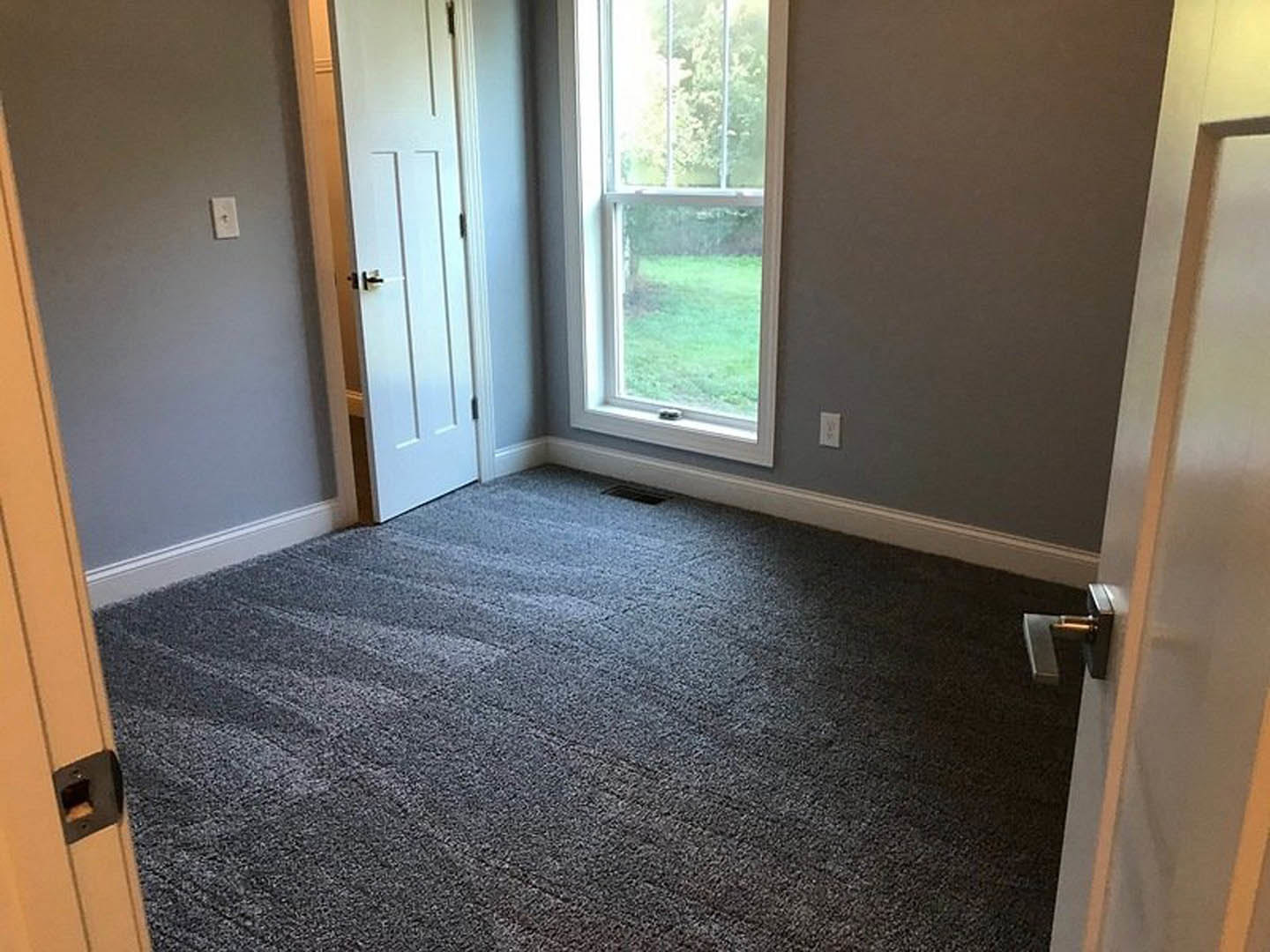 Carpeted room with white walls, wood door featuring a metal handle, rectangular window overlooking green grass, wall-mounted light switch, and ceiling vent