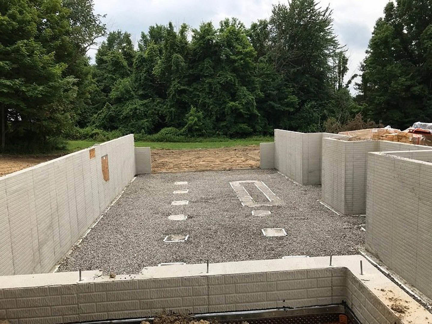 Gravel-covered building site with concrete foundation featuring square openings, surrounded by mature trees under a clear blue sky