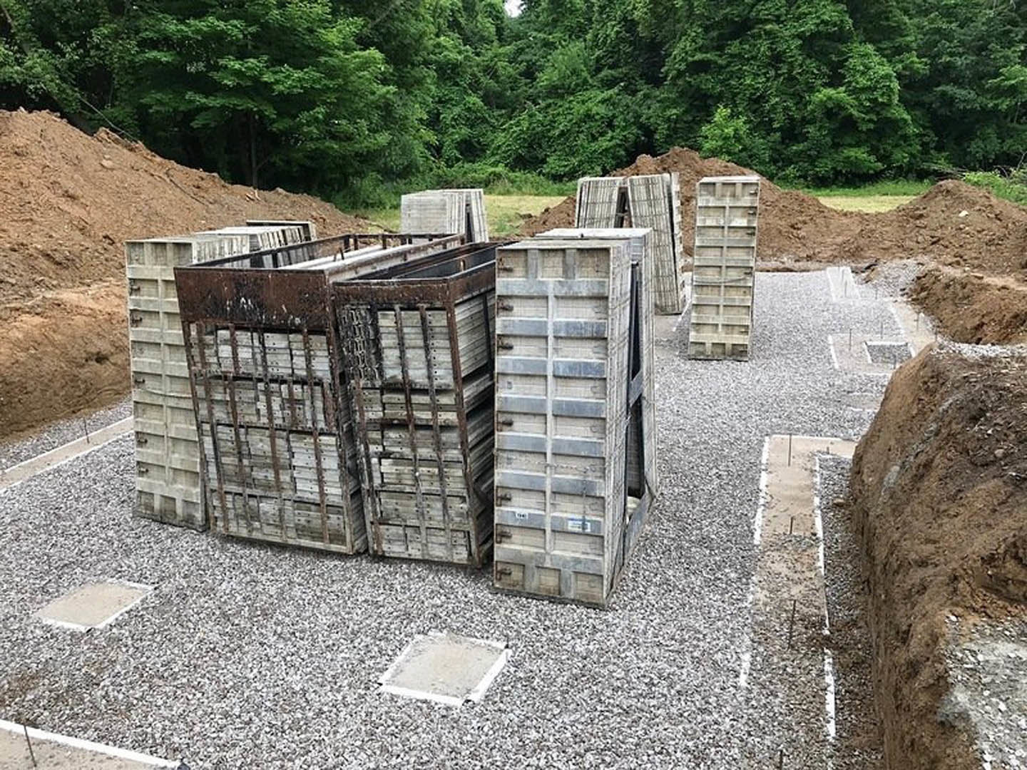 Stack of wooden pallets resting on gravel beside a metal gate, surrounded by trees and plants.