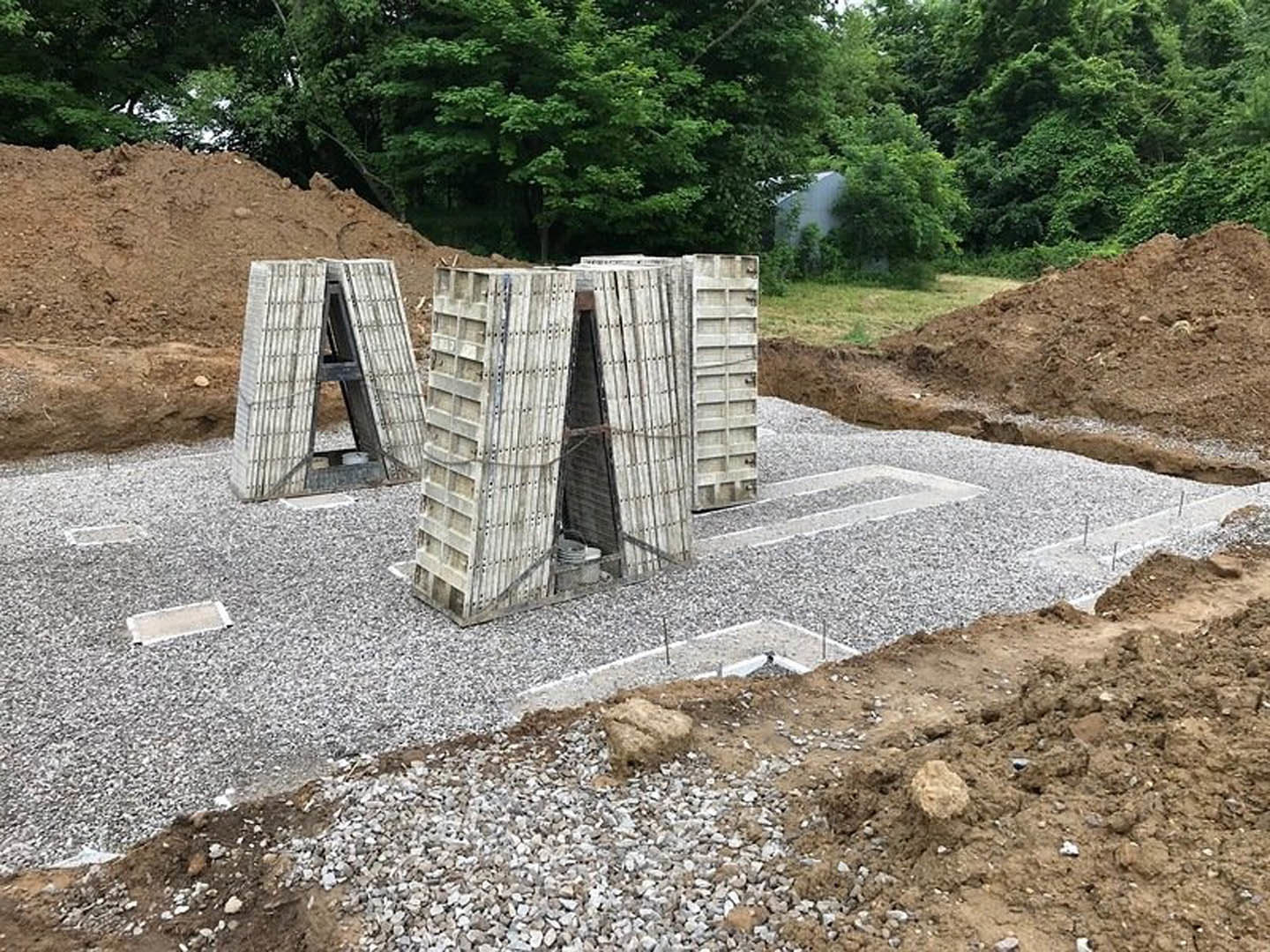 Triangular concrete structure with central opening surrounded by soil, rocks, and construction debris; leafy tree and wooden framing visible in background