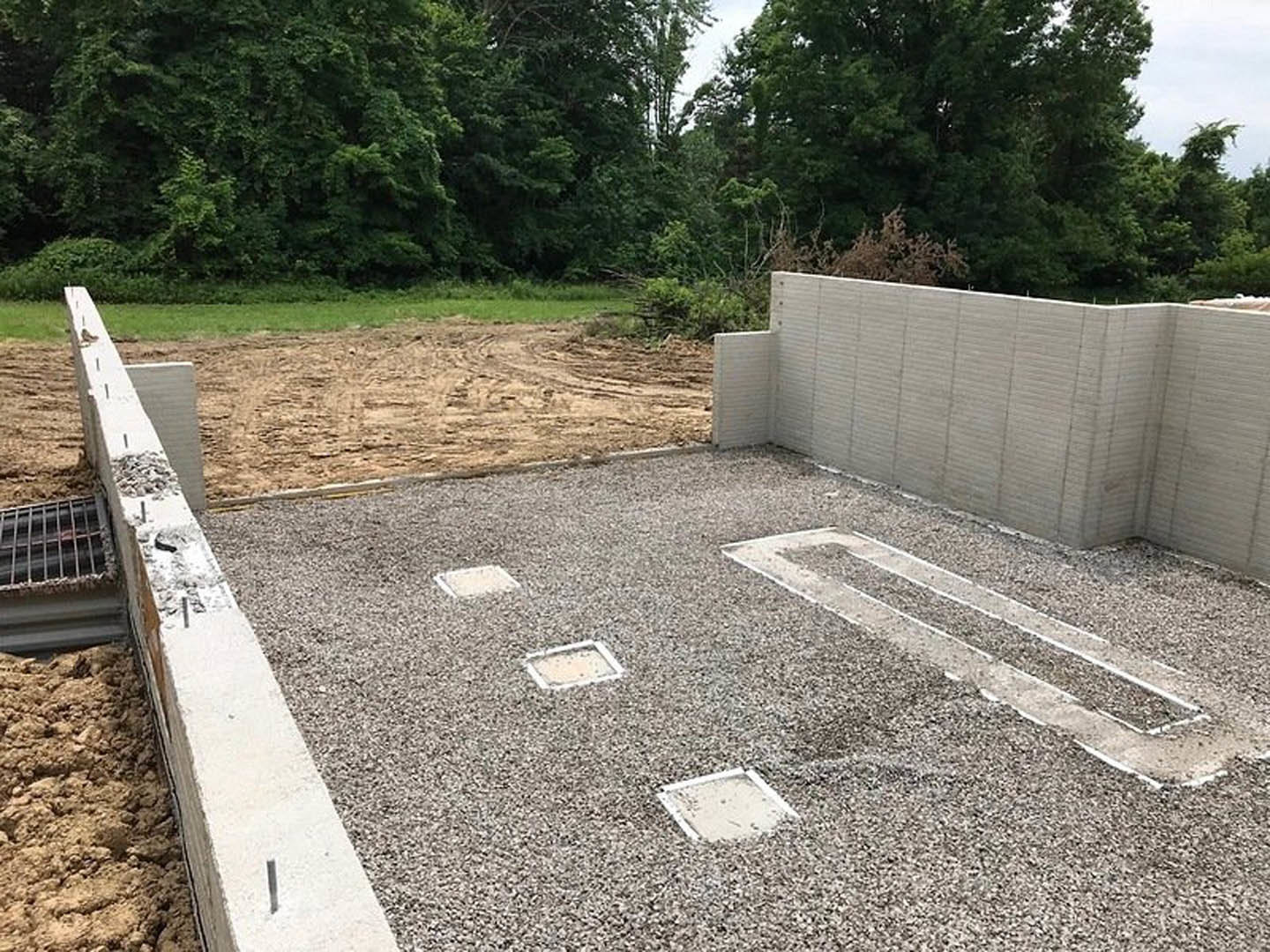 Concrete foundation with square holes, surrounded by gravel and dirt field, bordered by a brick wall and metal grate, with trees and plants visible in the background under a clear