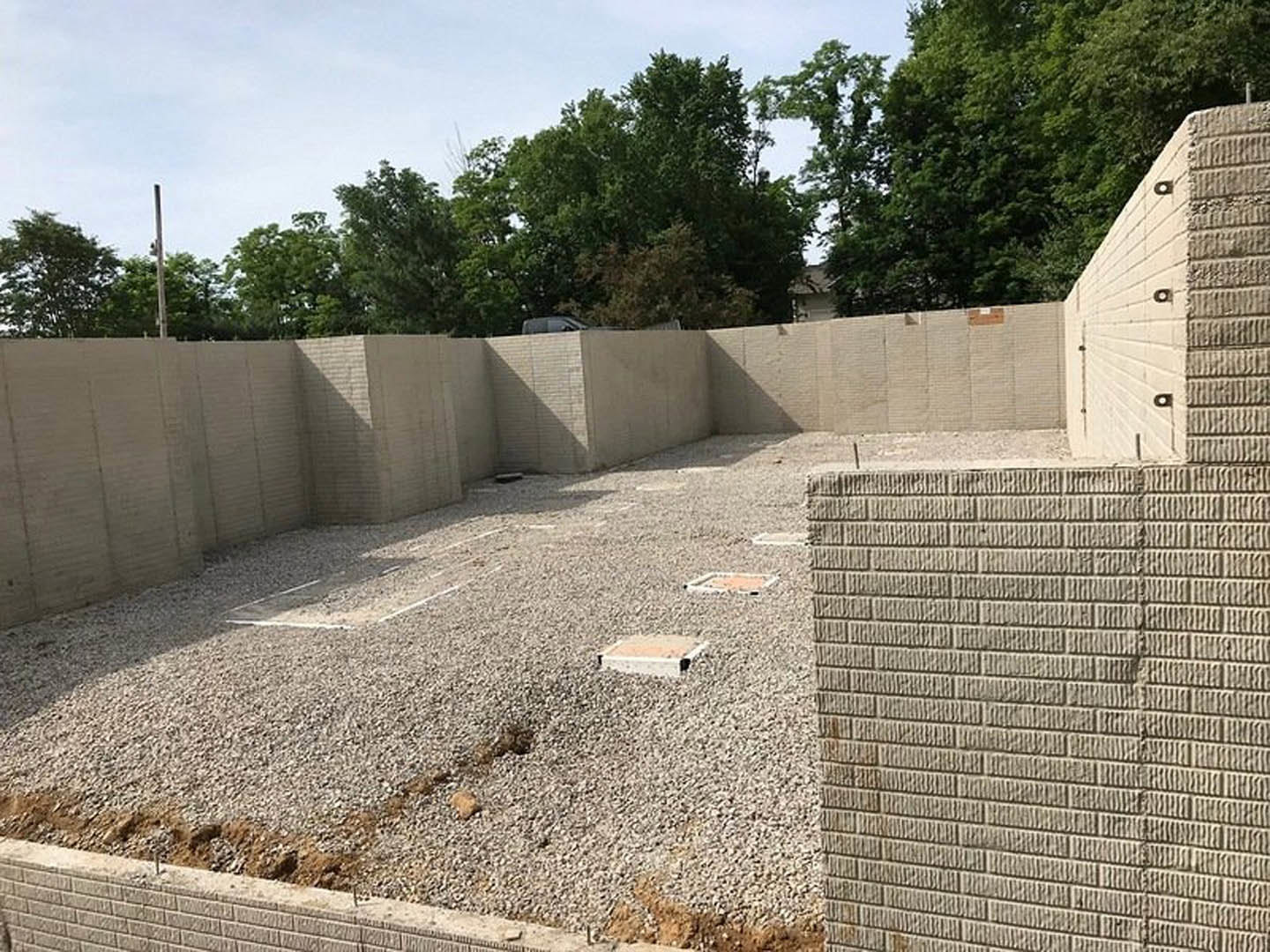 Partially built brick and stone exterior walls atop a gravel and concrete foundation, curb detail in foreground, leafy trees and blue sky in background, sunlight casting shadows on