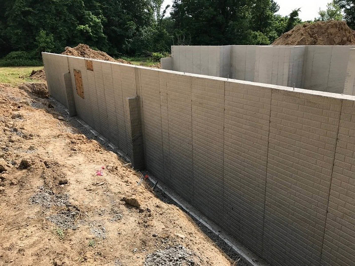 Concrete foundation wall with brown soil piled alongside, white and brown composite siding above, trees and sky visible in background