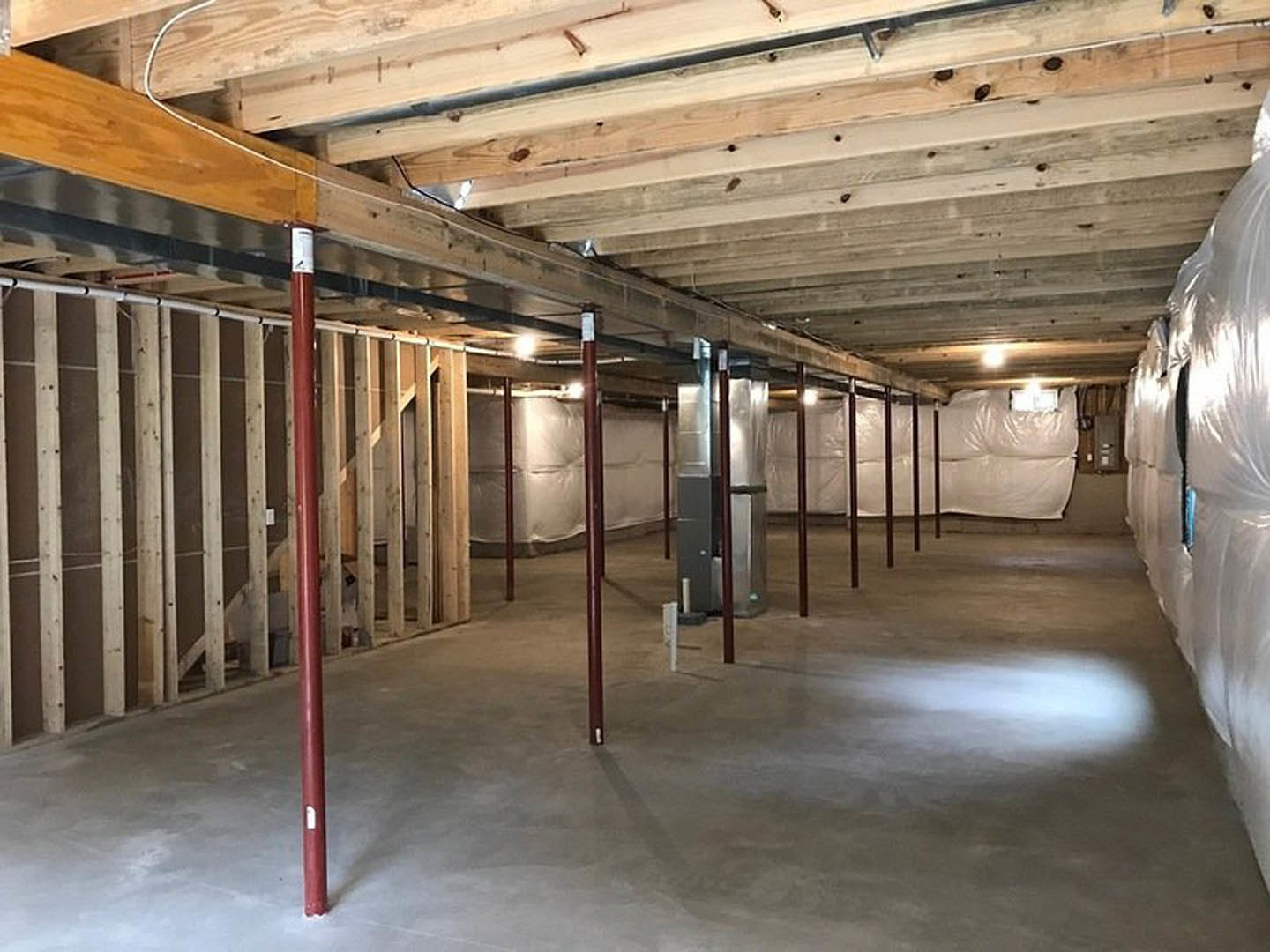 Wooden ceiling beams and exposed metal support poles above a concrete floor, unfinished walls with insulation and a white sheet, construction materials including stacked plastic