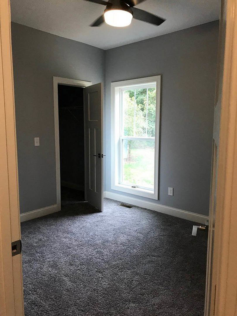 Carpeted room with white plaster walls, open door with silver handle, large window showing leafy trees, ceiling fan, recessed light, and floor vent.