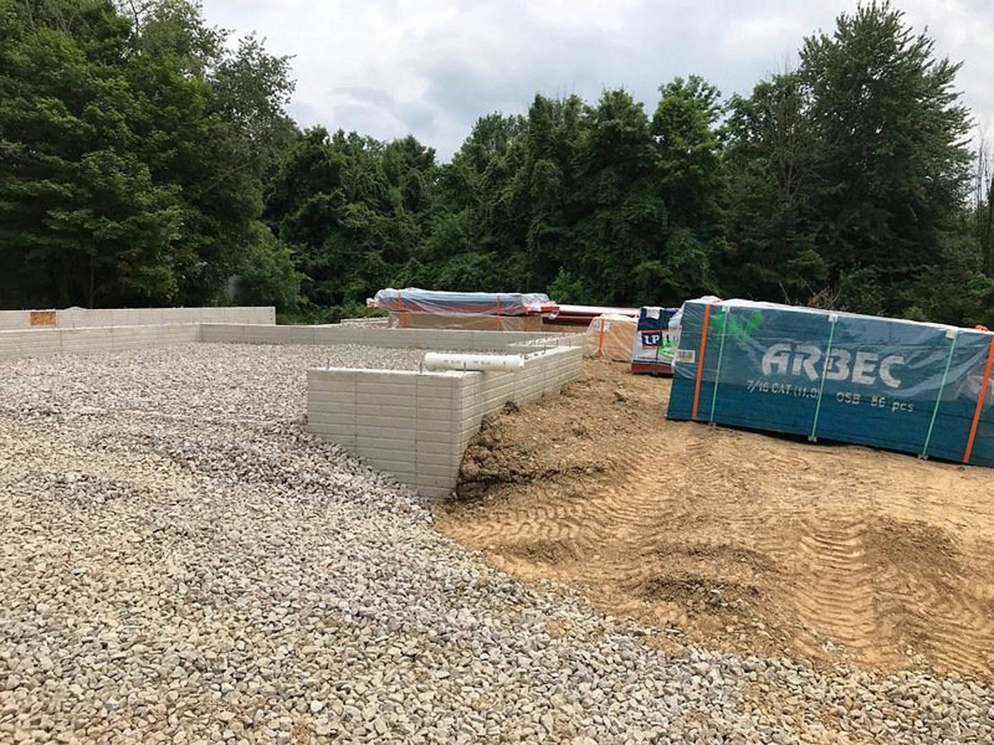 Gravel piles on bare soil beside a partially built foundation, blue tarp with white text, construction truck parked near wooded area under open sky