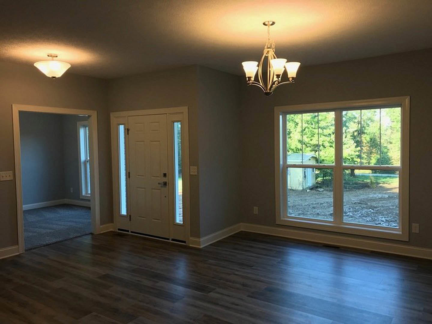 Dark wood flooring in a bright room with white walls, large windows showing trees outside, a white door with glass panels, and a close-up of a chandelier and modern light fixture
