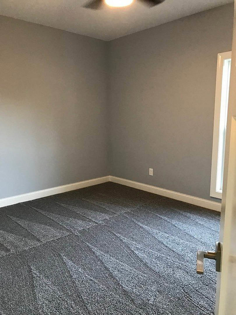 Grey carpeted bedroom with white walls, open door revealing hallway, ceiling fan overhead, large window letting in natural light, brushed metal door handle in foreground.