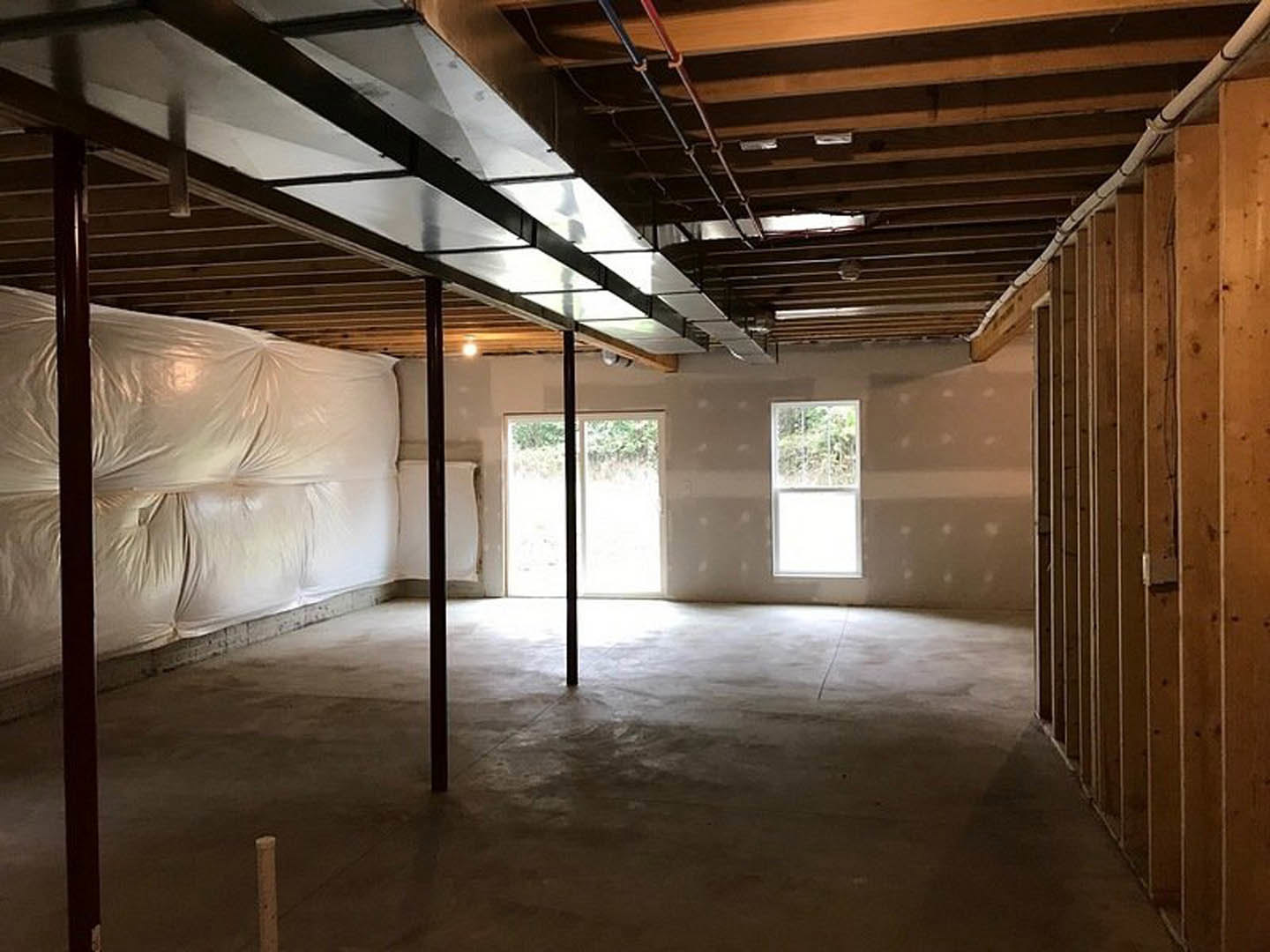 Basement room with exposed beams, plaster walls, white plastic sheet covering one wall, white-framed window, and close-up of a door.