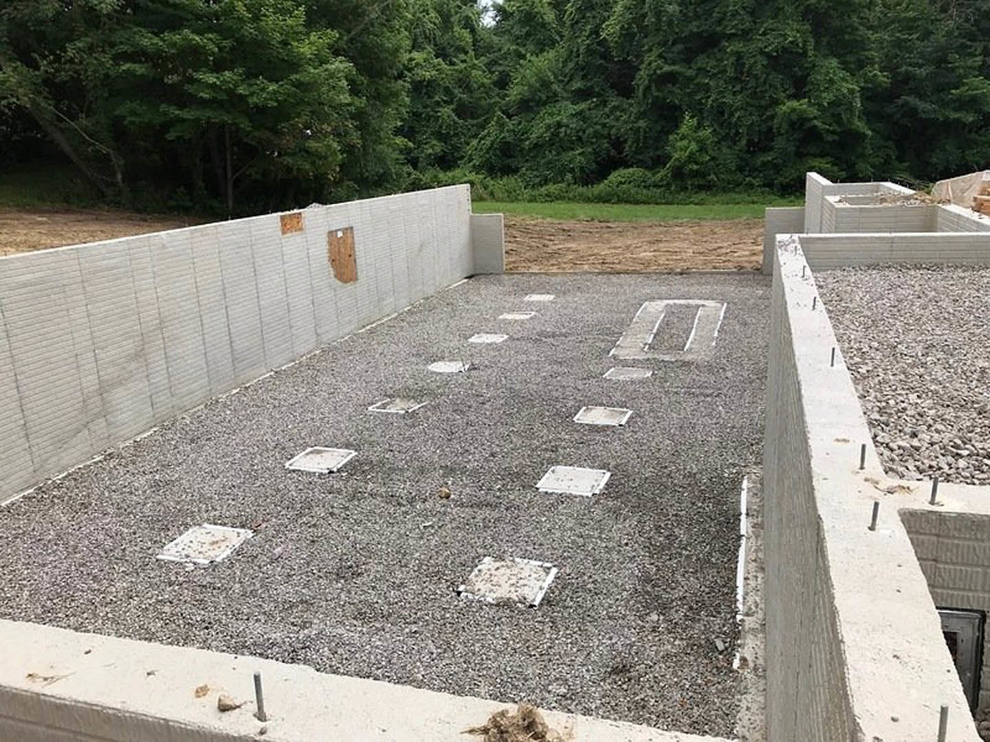 Concrete foundation with gravel fill and square concrete blocks, surrounded by green trees and plants in a wooded outdoor setting.