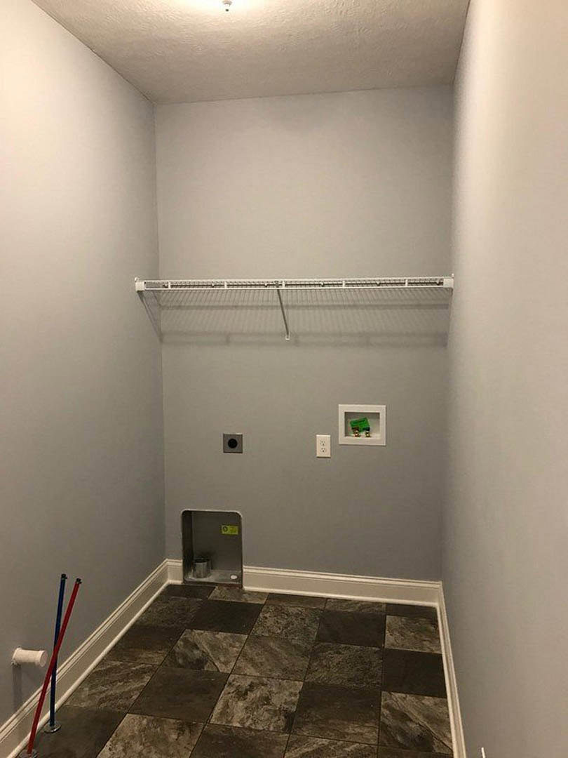 White plaster wall with floating metal shelf, tile flooring, red and blue broomsticks leaning against the wall, green object in a white frame, metal bin in a rectangular box.