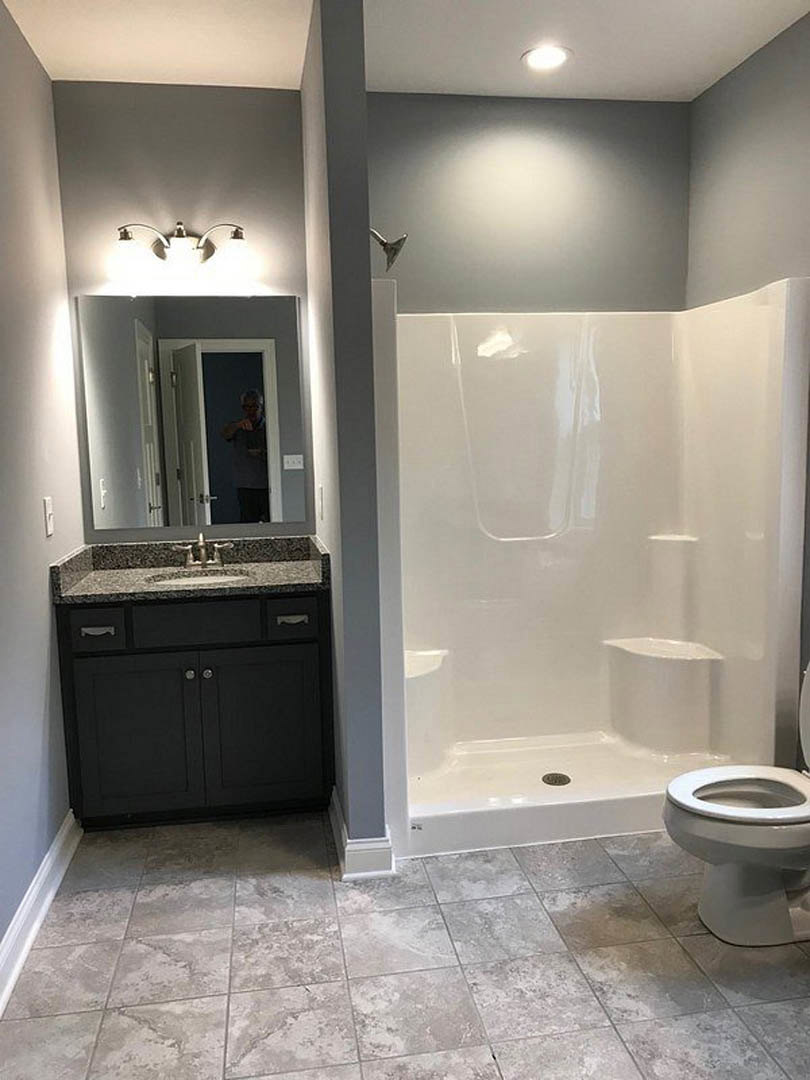 Modern bathroom featuring a white tile shower, white walls, a ceramic sink, and a close-up of gray tile flooring; a man stands in the doorway holding a black object near a white