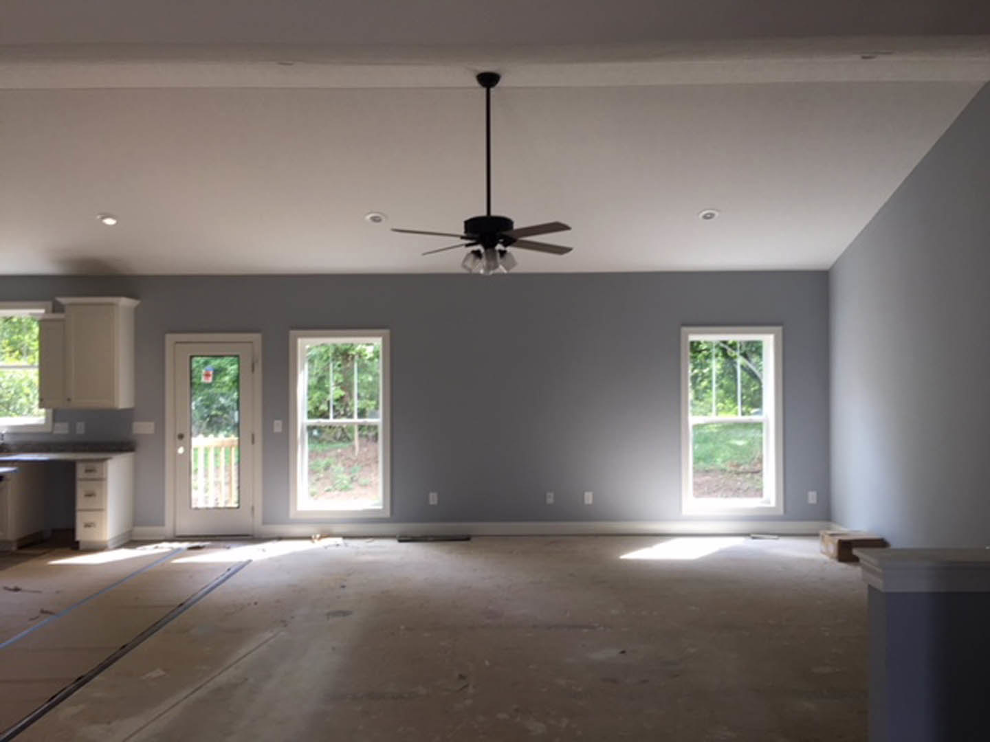 Ceiling fan mounted on white ceiling, large windows with views of green lawn, glass-paneled door, hardwood flooring, neutral walls, small table in foreground