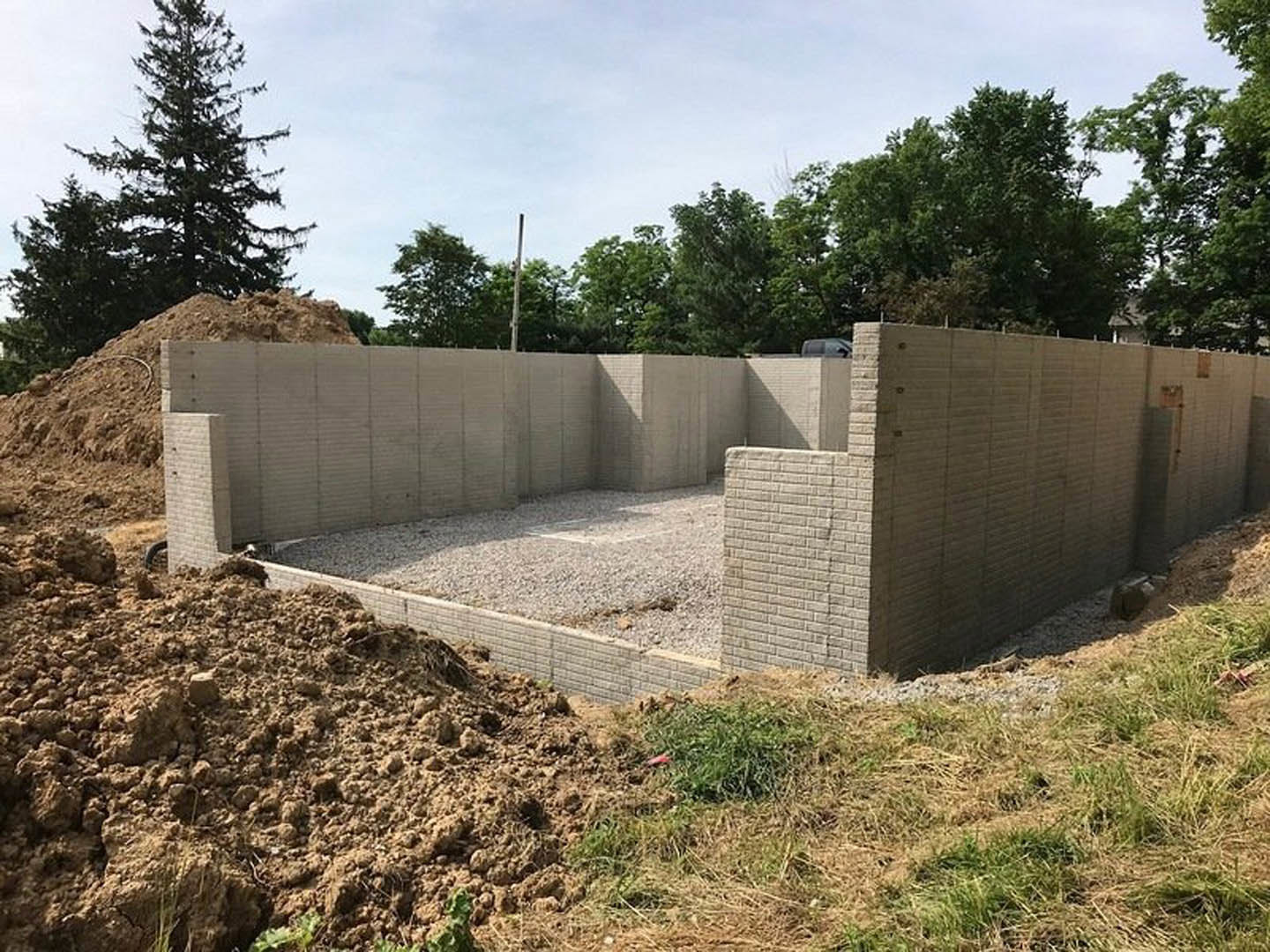 Concrete foundation and brick wall under construction, gravel and pile of dirt in foreground, group of trees and cloudy sky in background