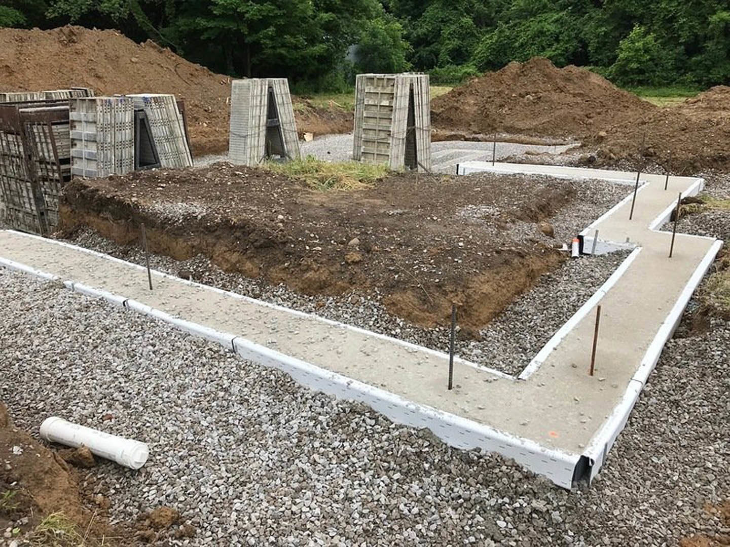 Concrete foundation pillars surrounded by dirt and gravel, wire mesh reinforcement, white pipe laying on the ground, exposed soil and rocks, tree and plants in the background