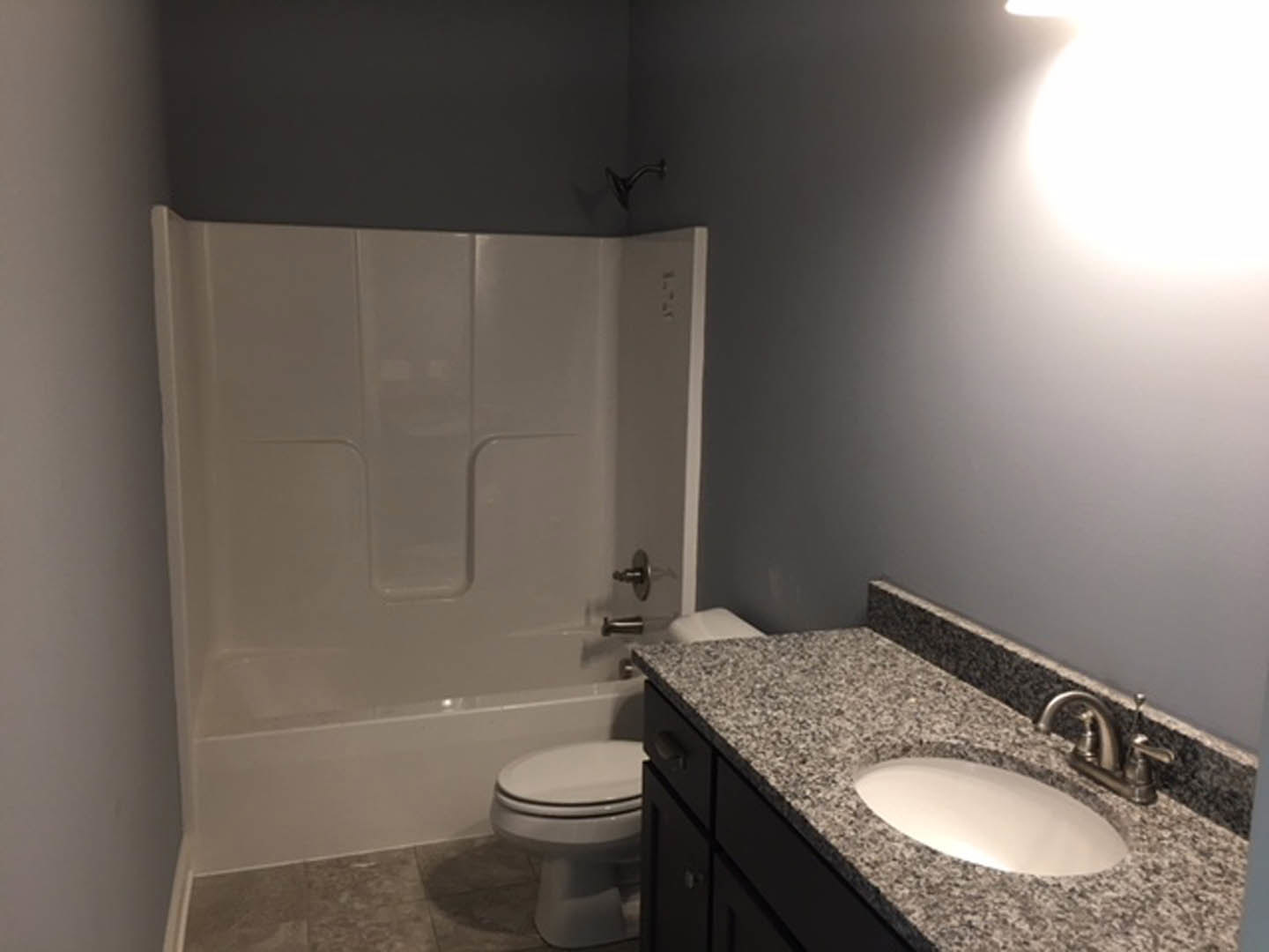 Granite countertop with white oval sink, chrome faucet, glass-enclosed shower, white toilet, large wall mirror, and light gray tile walls