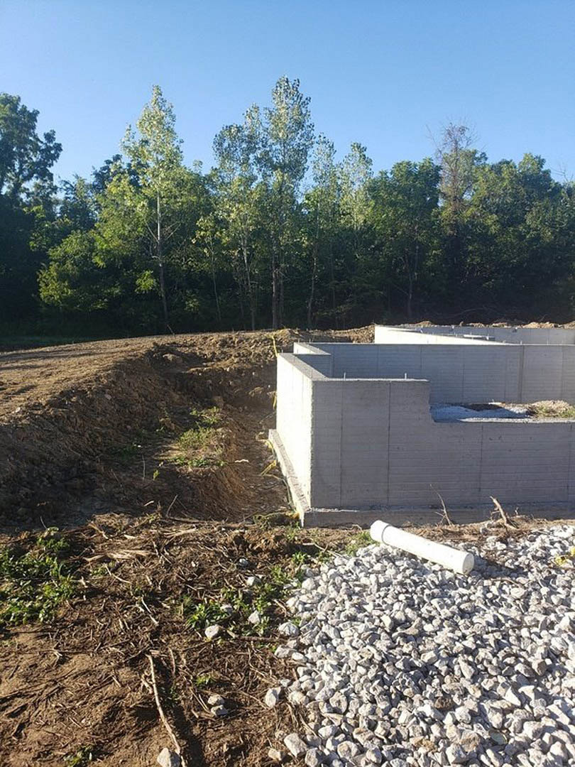 Concrete foundation wall with white wood beam resting on rocks, white pipe lying nearby, dirt ground scattered with sticks and rocks, surrounded by trees and plants under open sky.