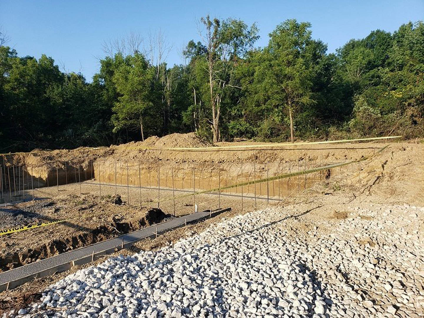 Dirt foundation bordered by trees, gravel road with a pile of rocks, leafless tree in the background