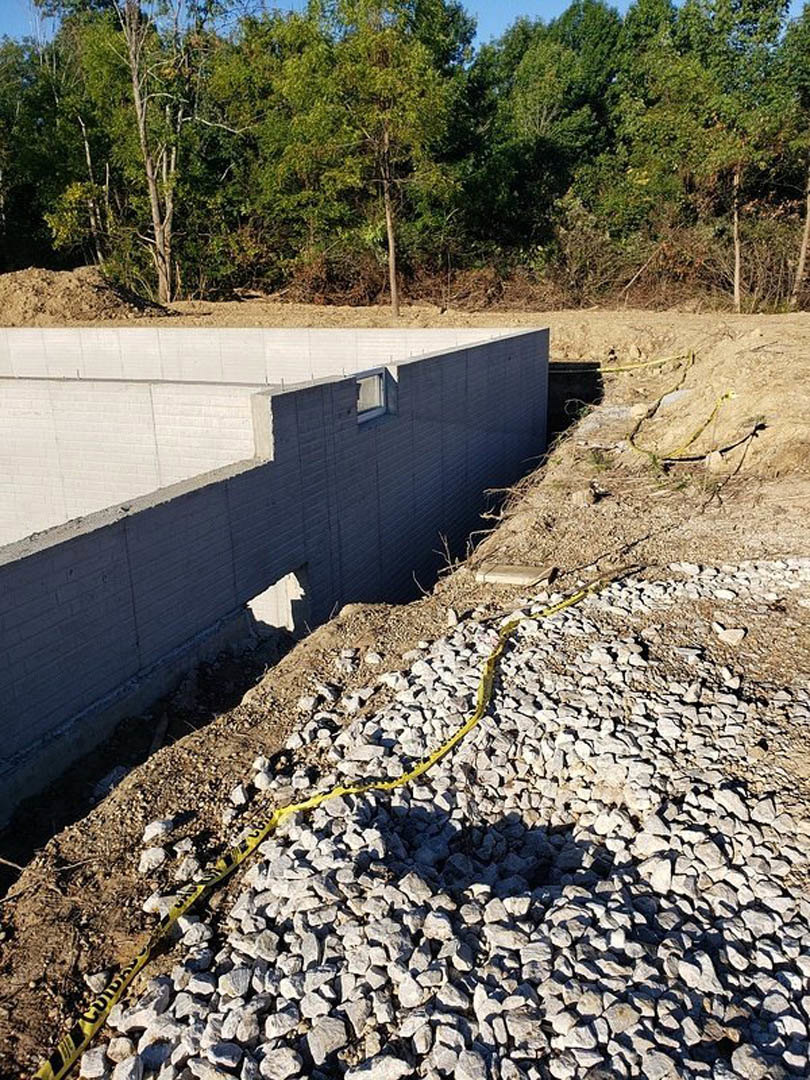 Concrete foundation with yellow hose, gravel and rocks scattered nearby, tree in forested background, unfinished concrete wall with visible hole