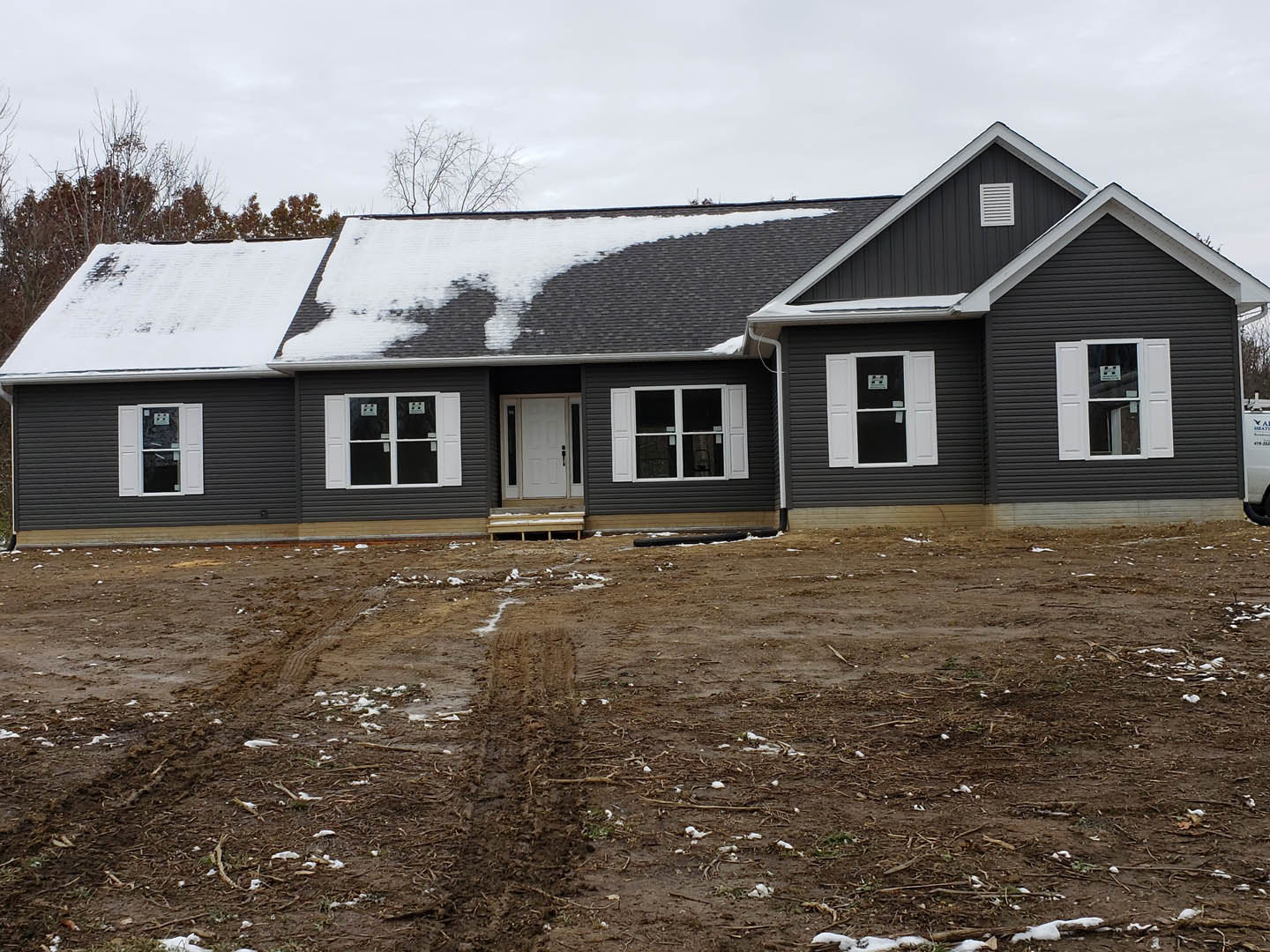 Two-story house with white siding, snow covering the roof and ground, tire tracks in muddy driveway, white-framed windows including one with a sign, white door with glass panel