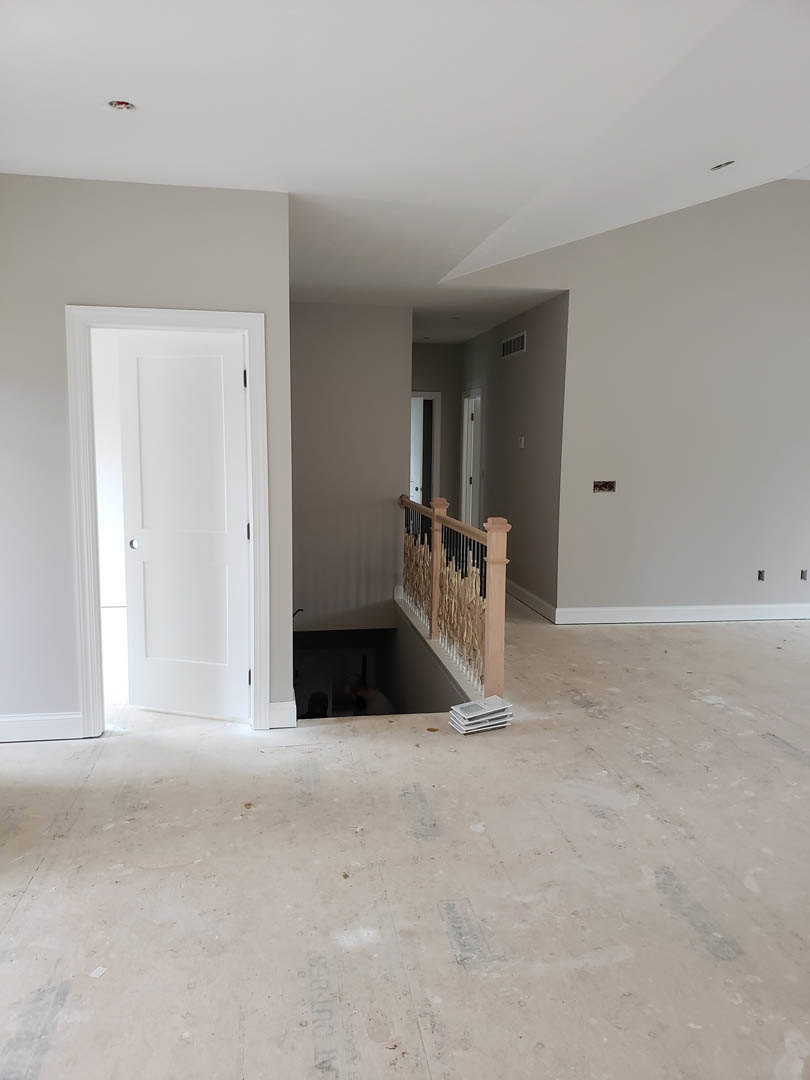 Staircase with wooden railing leading to unfinished basement, white paneled door with round knob, plaster walls, light-colored flooring, recessed ceiling.